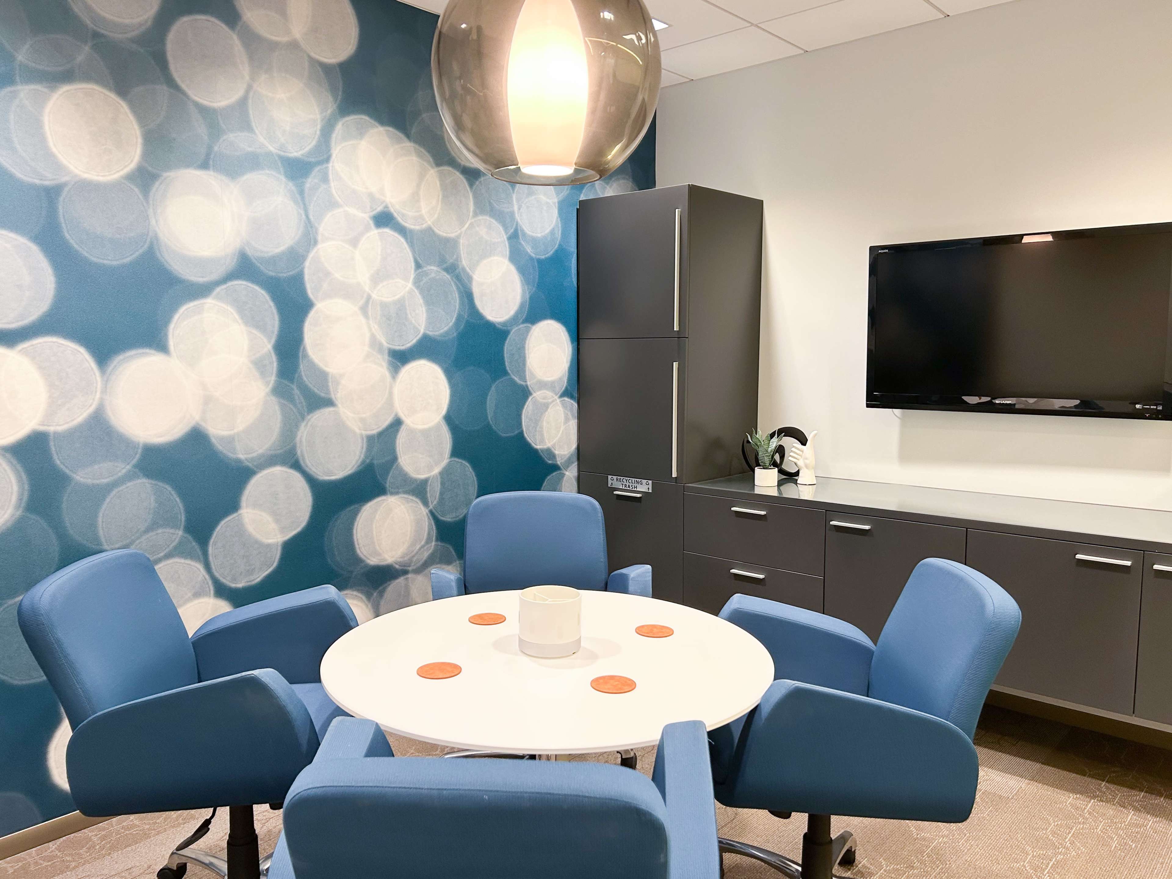 The image shows a conference room with a round table surrounded by four blue chairs, a television mounted on the wall, and a decorative backdrop featuring soft, illuminated dots.