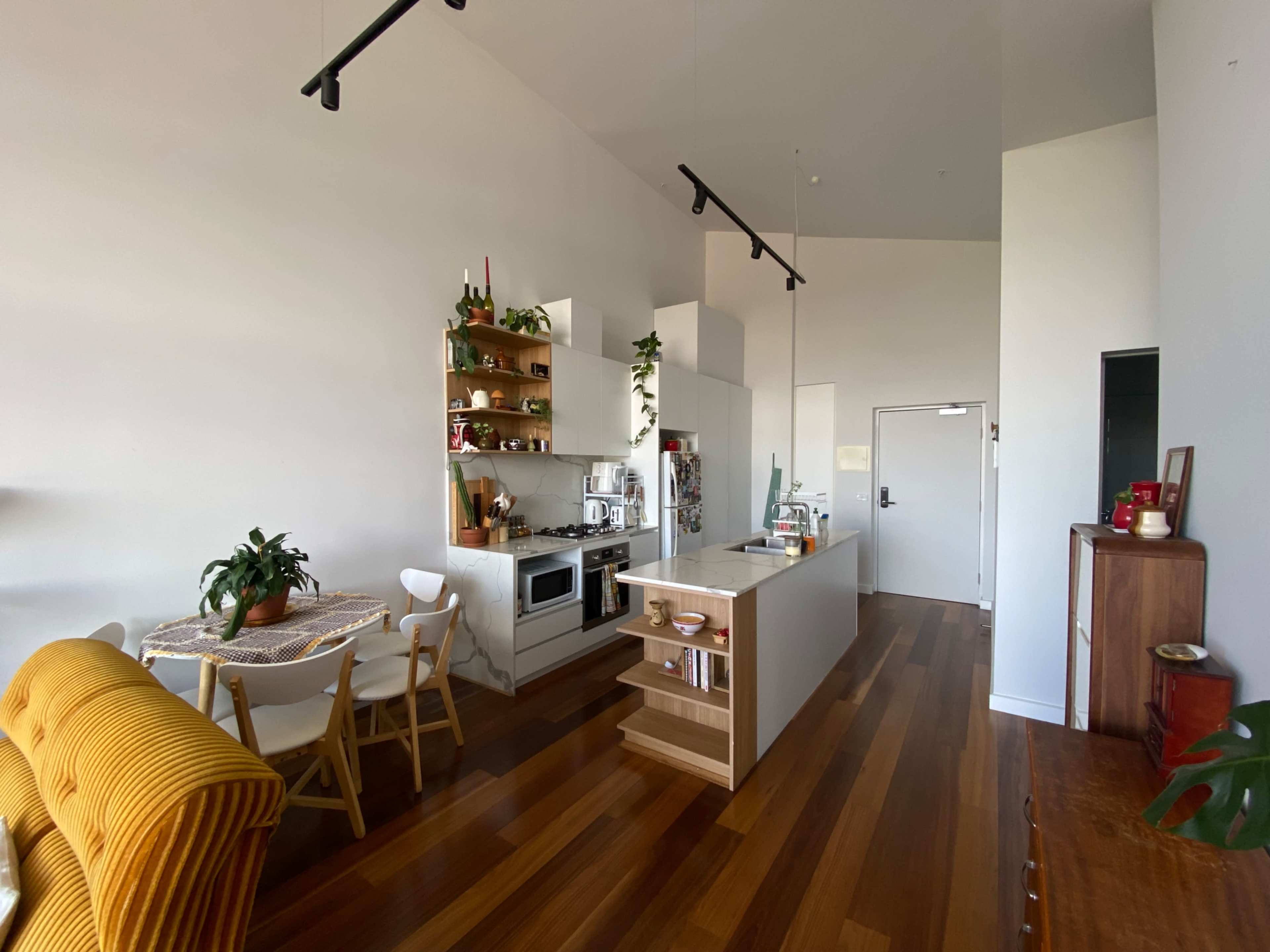 The image shows a modern kitchen and dining area featuring an island counter, wooden floors, and a dining table with chairs, all complemented by plants and open shelving.