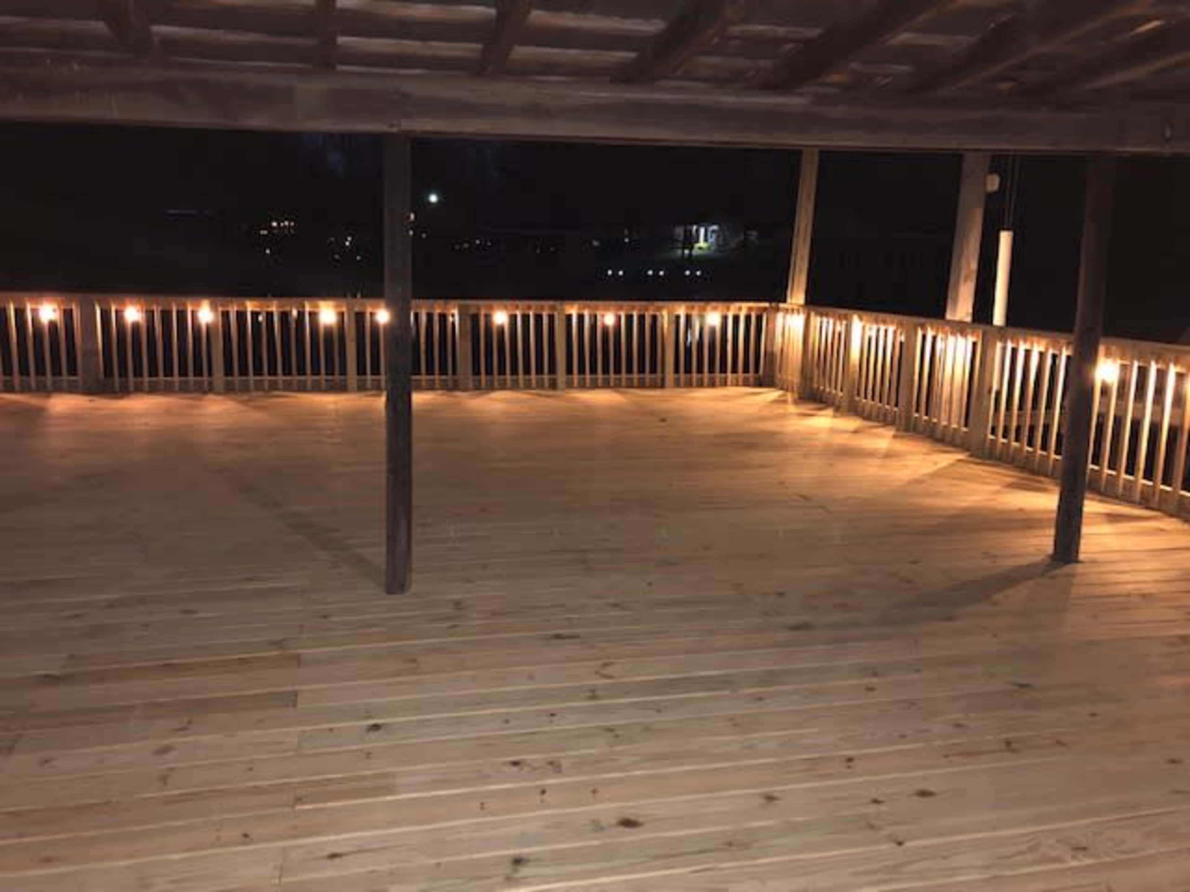 A wooden deck is illuminated by string lights, with an empty, spacious area and a railing around the perimeter.