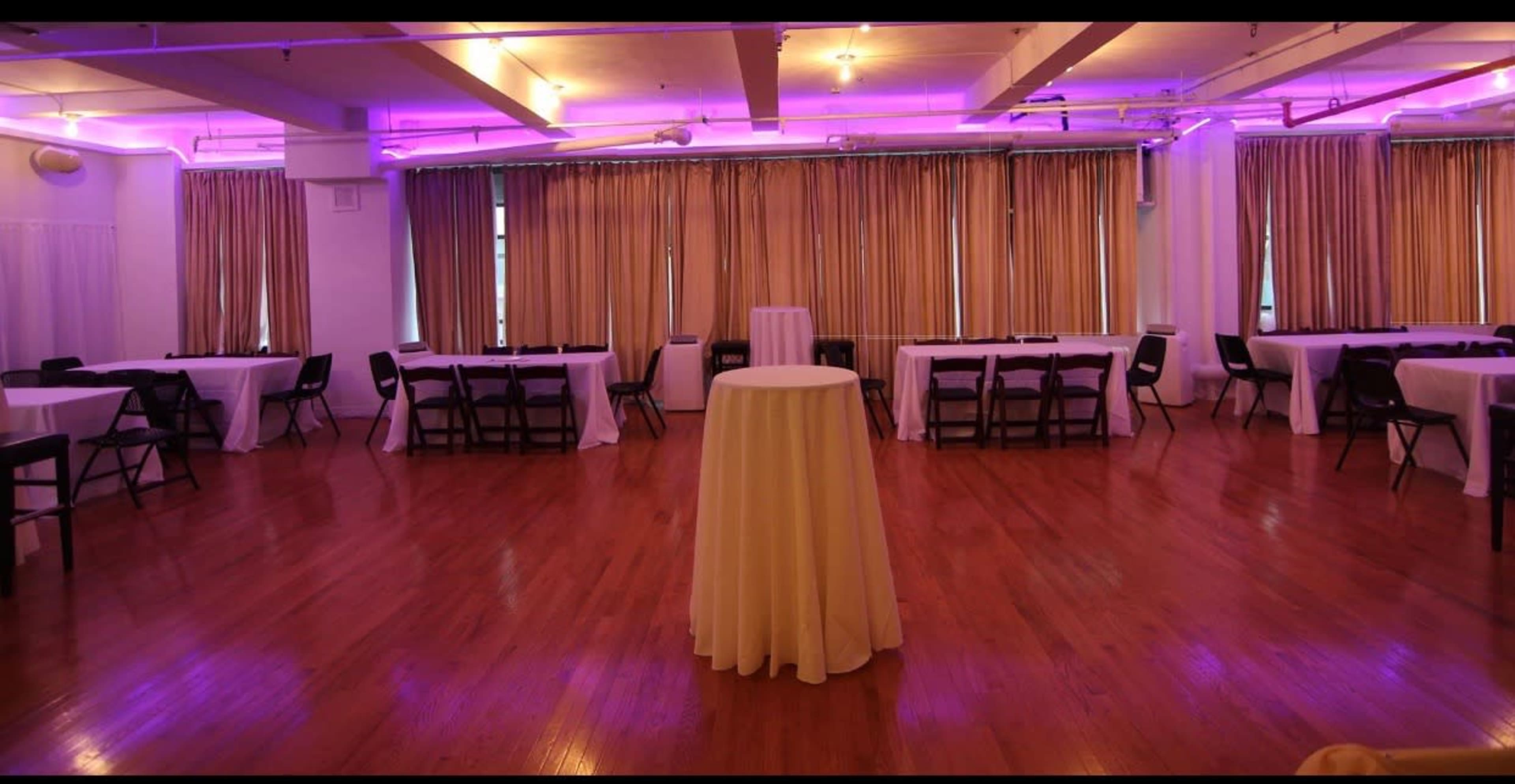 An empty event space with tables covered in white tablecloths and purple lighting illuminating the room.