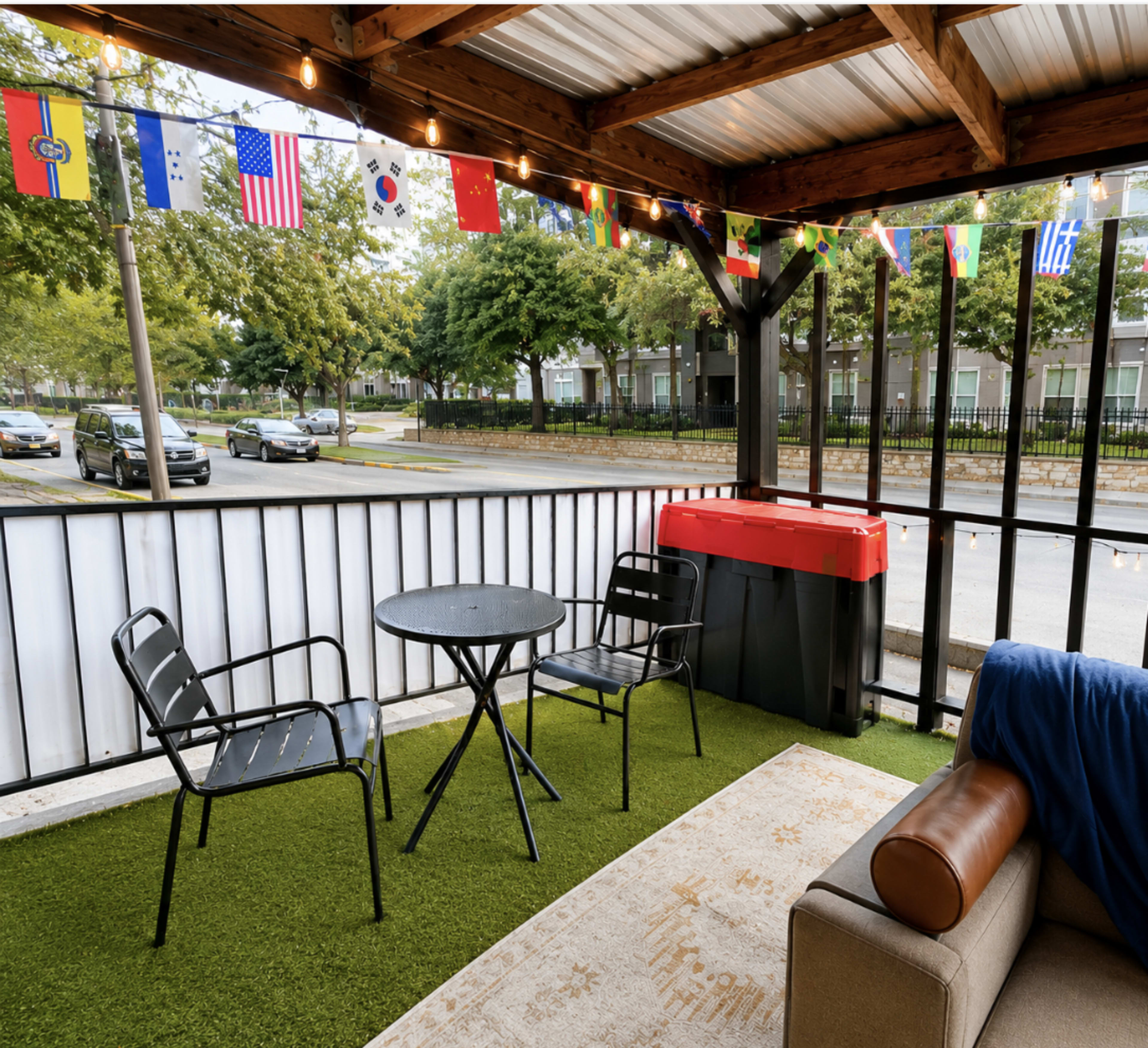 The image shows a covered outdoor patio with a small table and two chairs, decorated with string lights and flags from various countries.