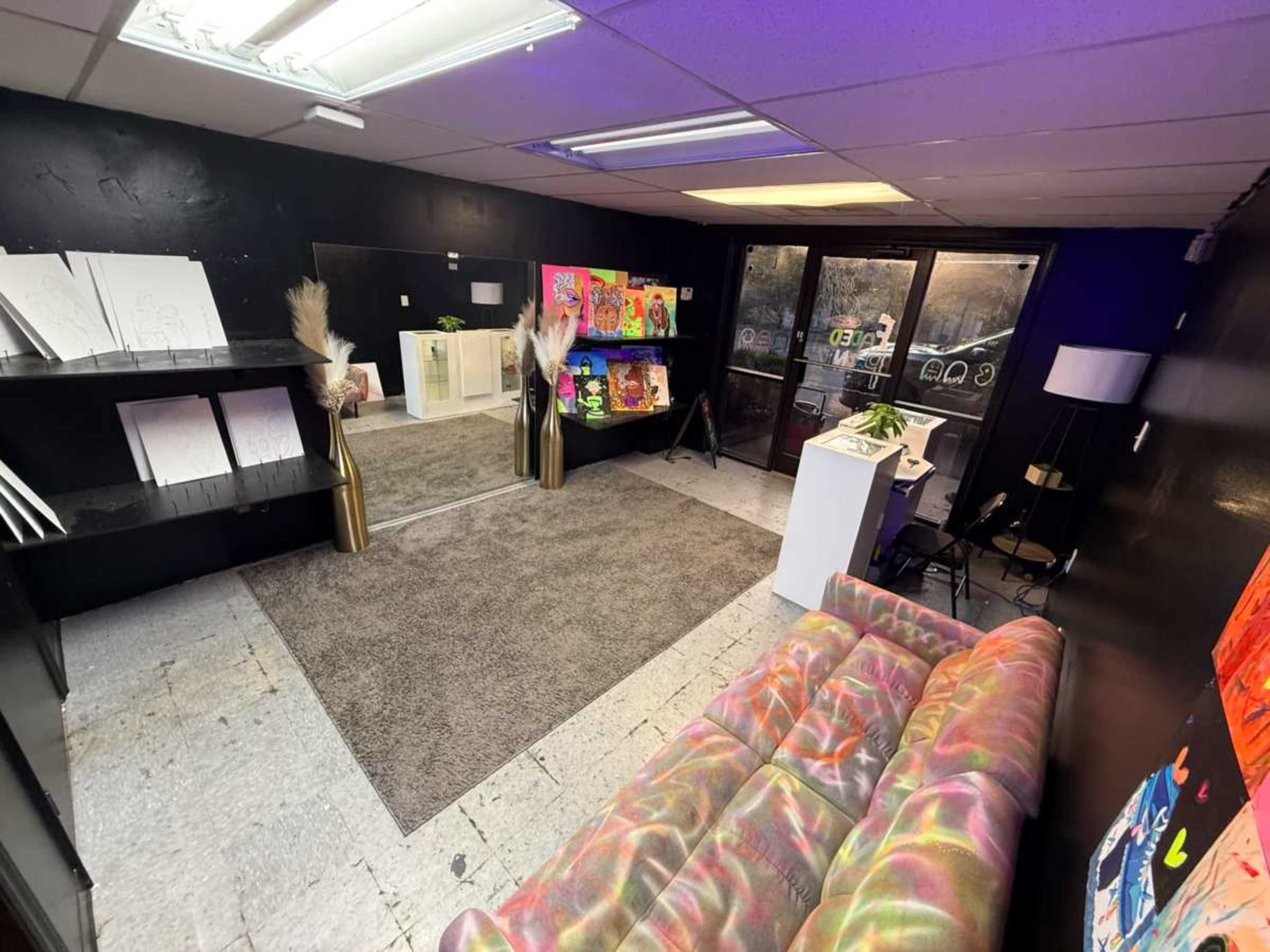The image shows an art gallery interior featuring a colorful sofa, shelves with artworks, and large glass doors.