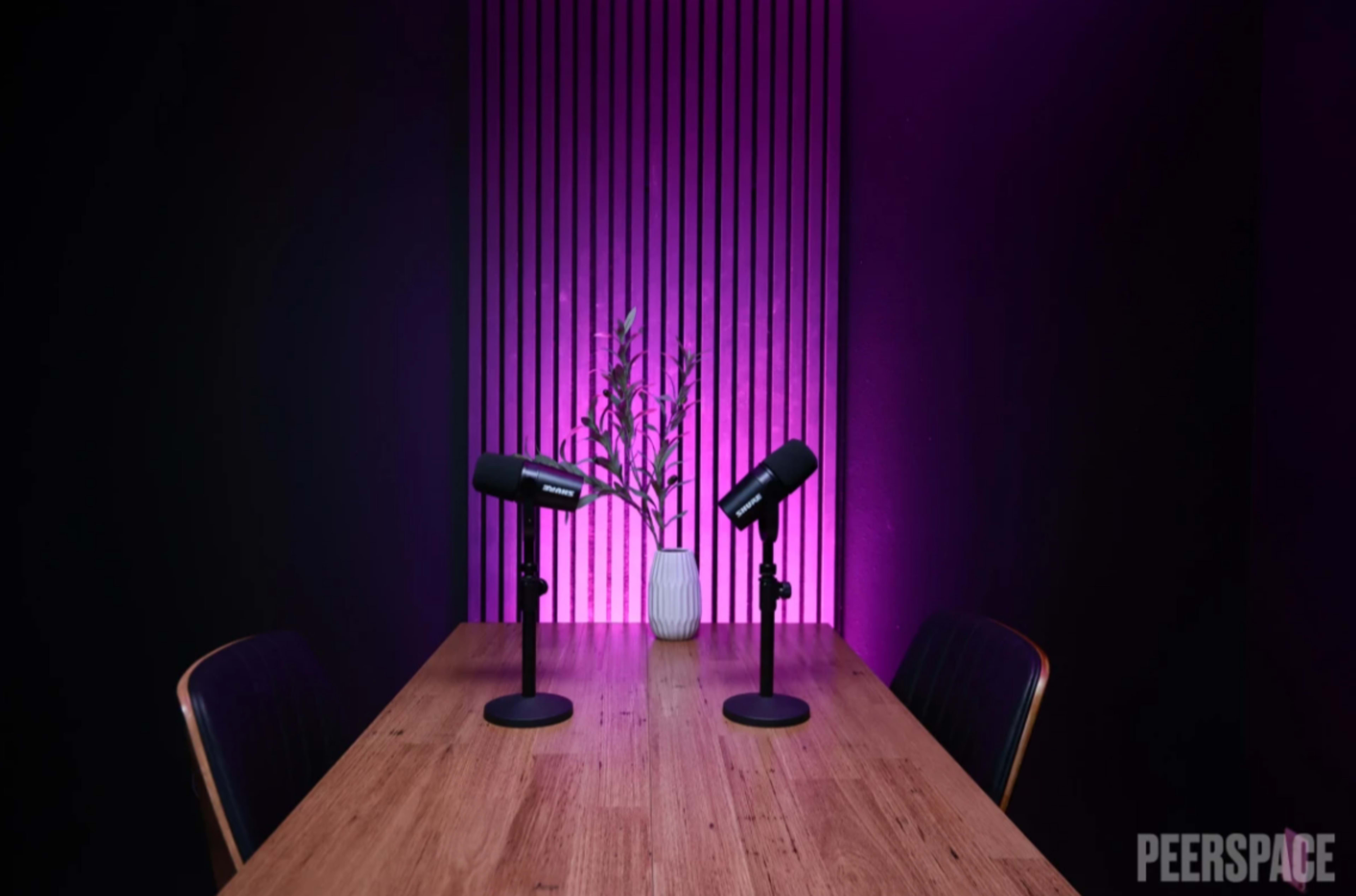 A wooden table with two microphones stands in front of a backdrop featuring vertical stripes lit in purple, with a vase of flowers in the center.