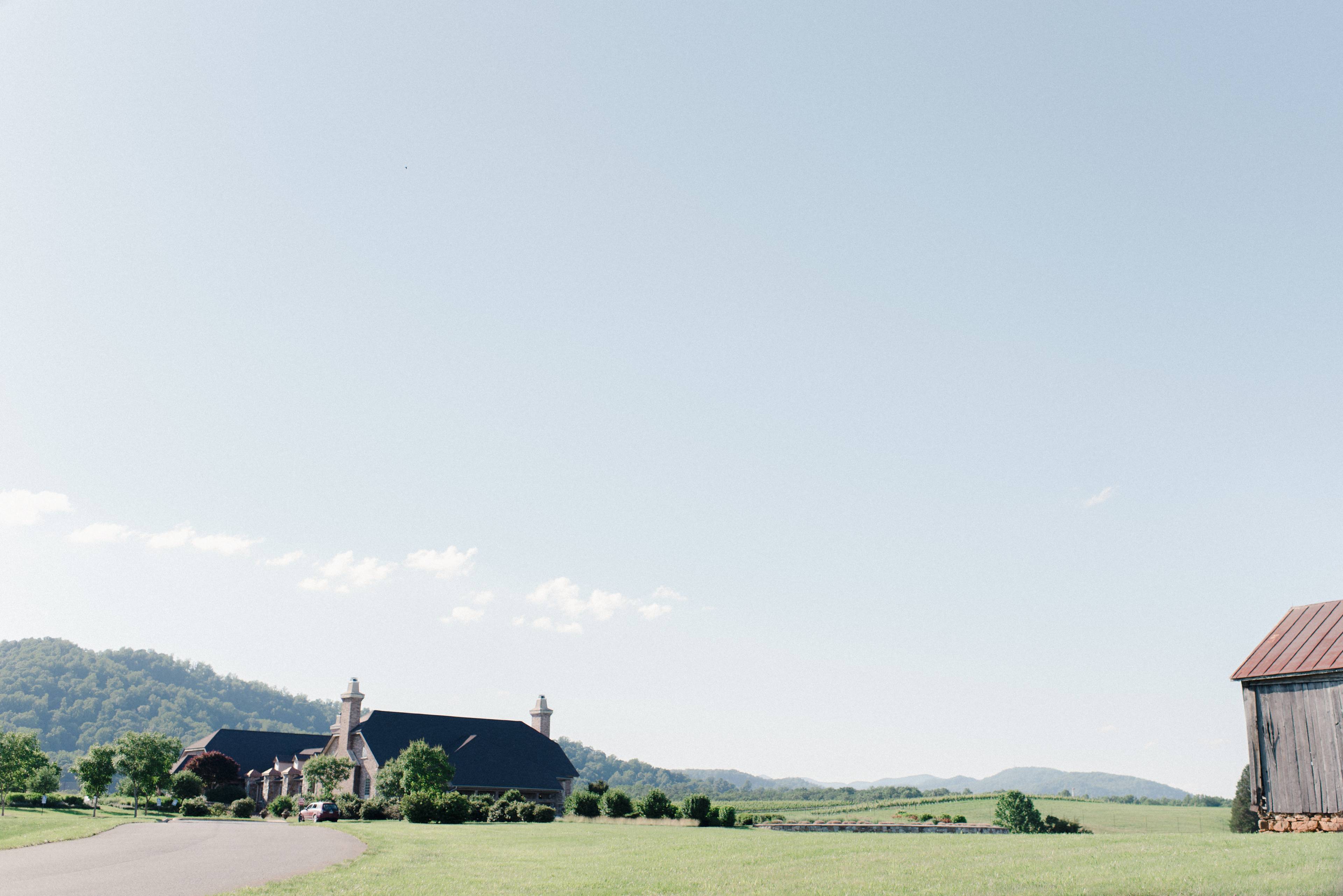 A single-story building with a dark roof stands alongside a road, surrounded by green fields and rolling hills under a clear sky.