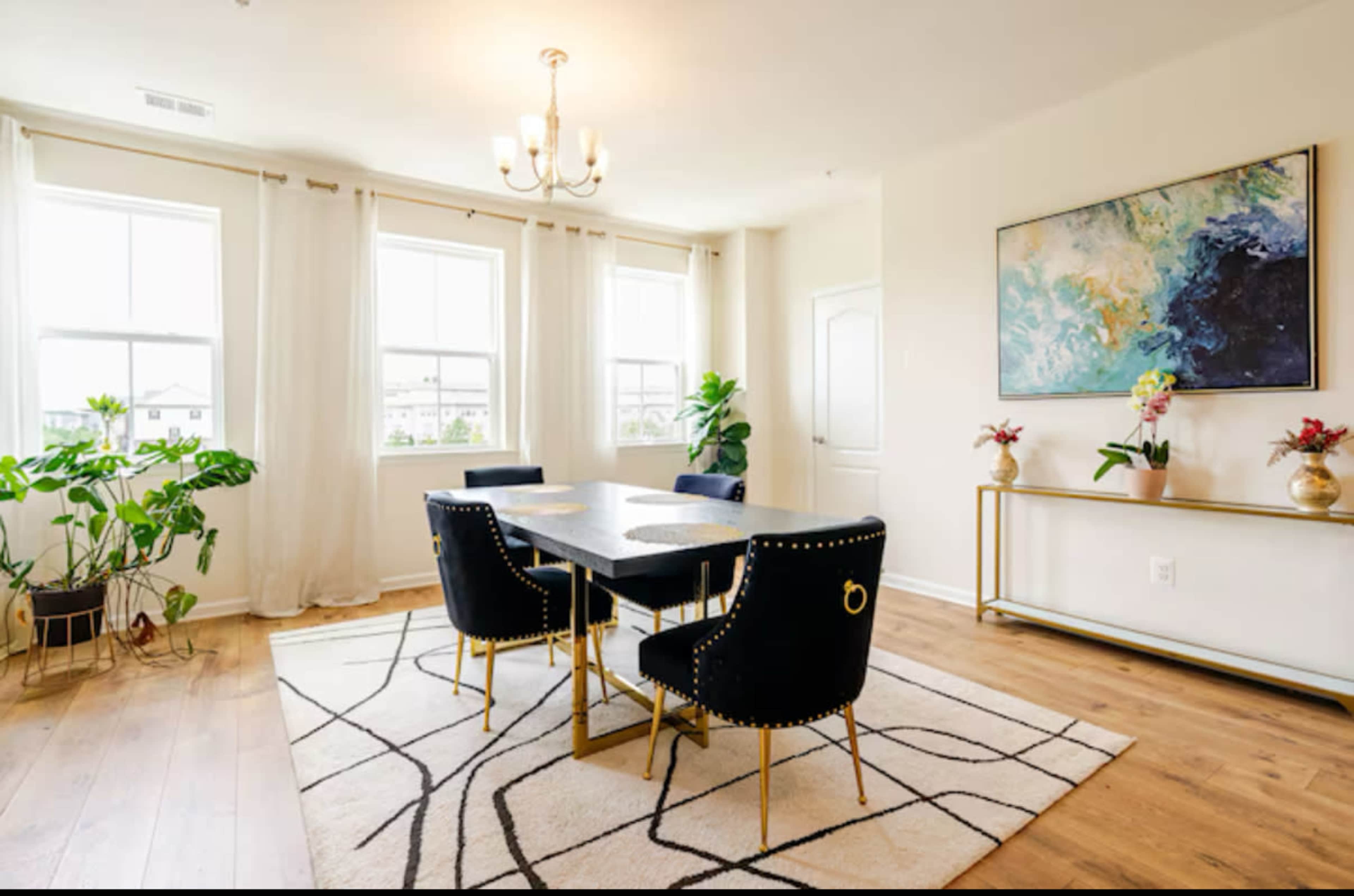 A dining area features a rectangular table surrounded by four black chairs, with large windows allowing natural light and potted plants adding greenery.