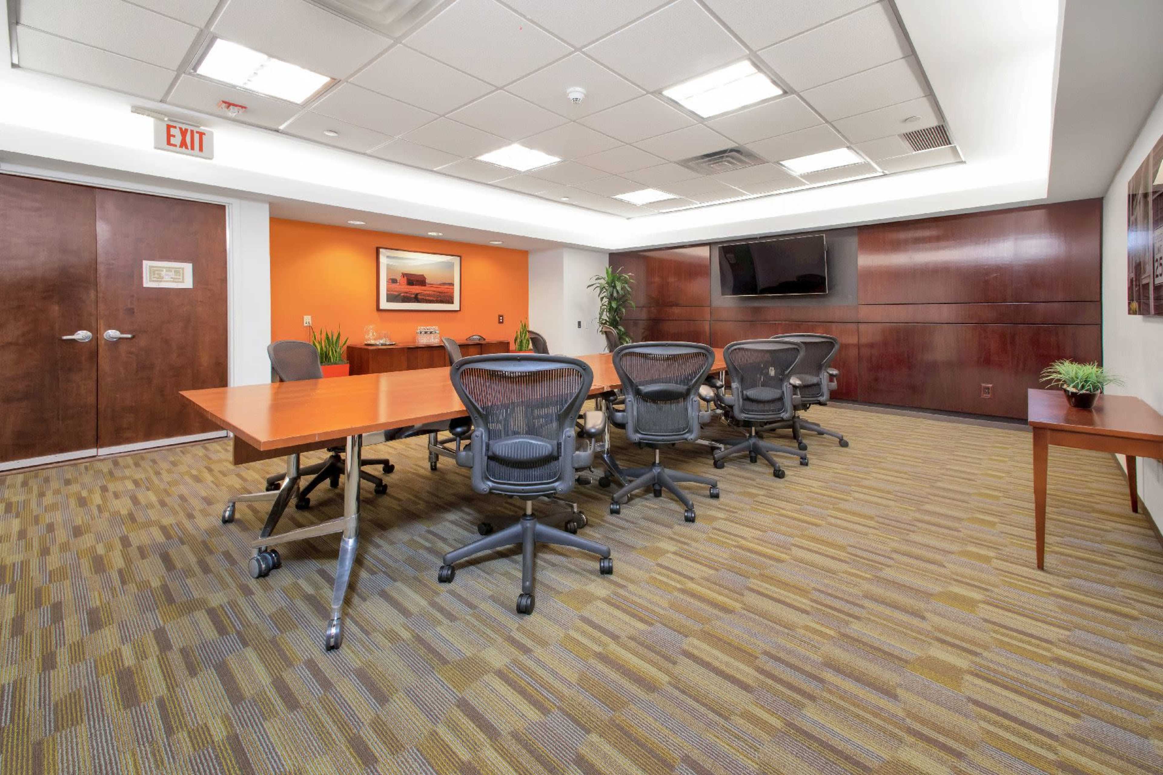 A conference room features a long wooden table surrounded by rolling office chairs, with an orange accent wall and a large television mounted on the wall.