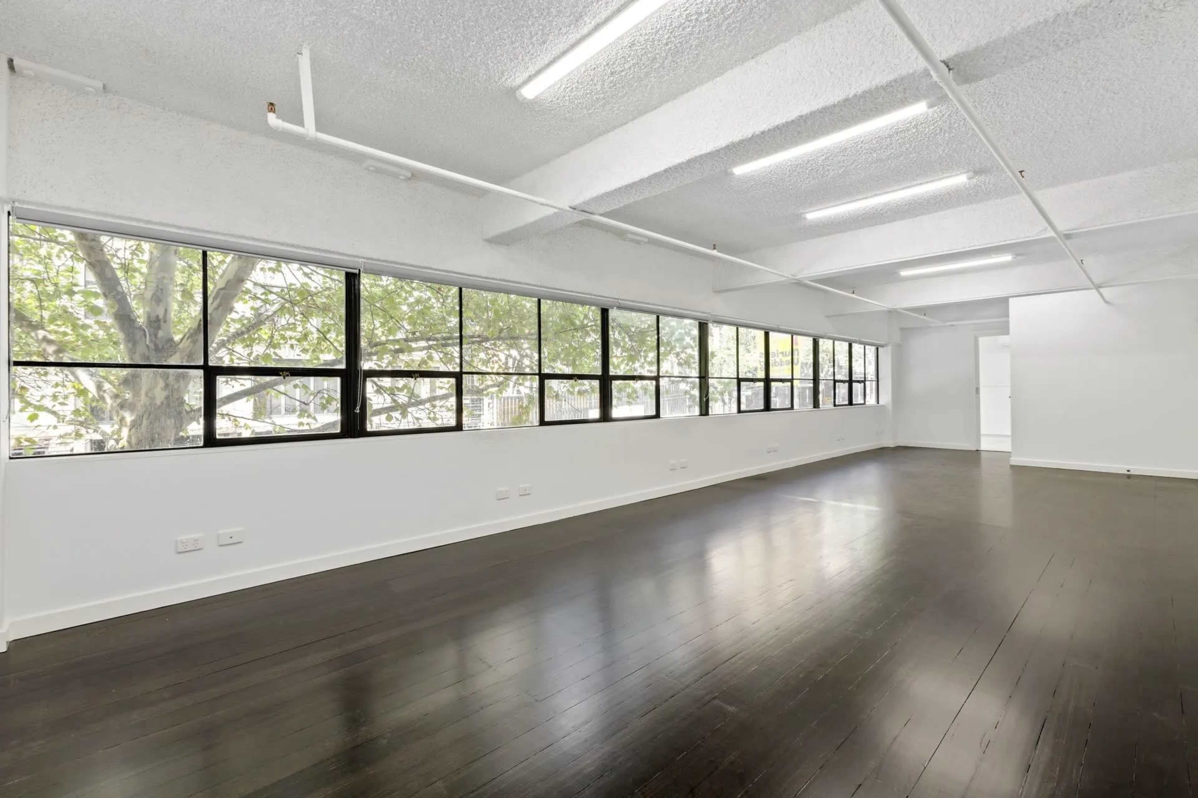 The image shows an empty room with dark wooden flooring and large windows providing natural light.