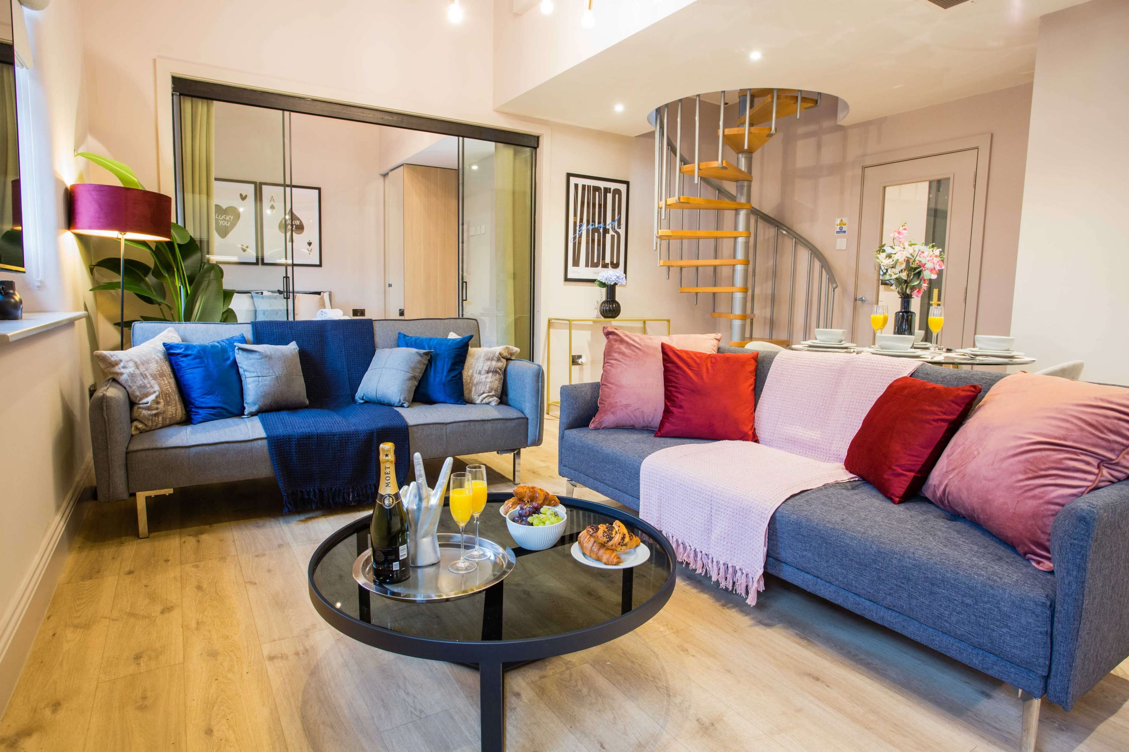 A modern living room features a cozy seating area with two sofas adorned with colorful cushions, a round coffee table set with snacks and drinks, and a spiral staircase leading to a lofted space.