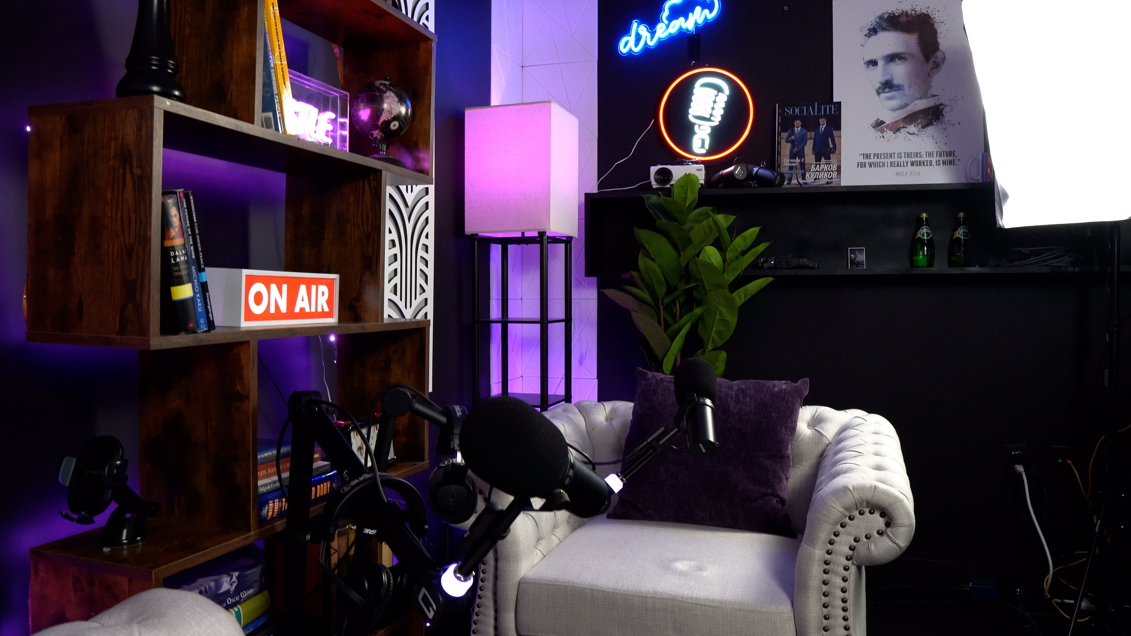 A cozy podcast recording setup featuring a white tufted armchair, a bookshelf filled with books, neon lighting, and a microphone on display.