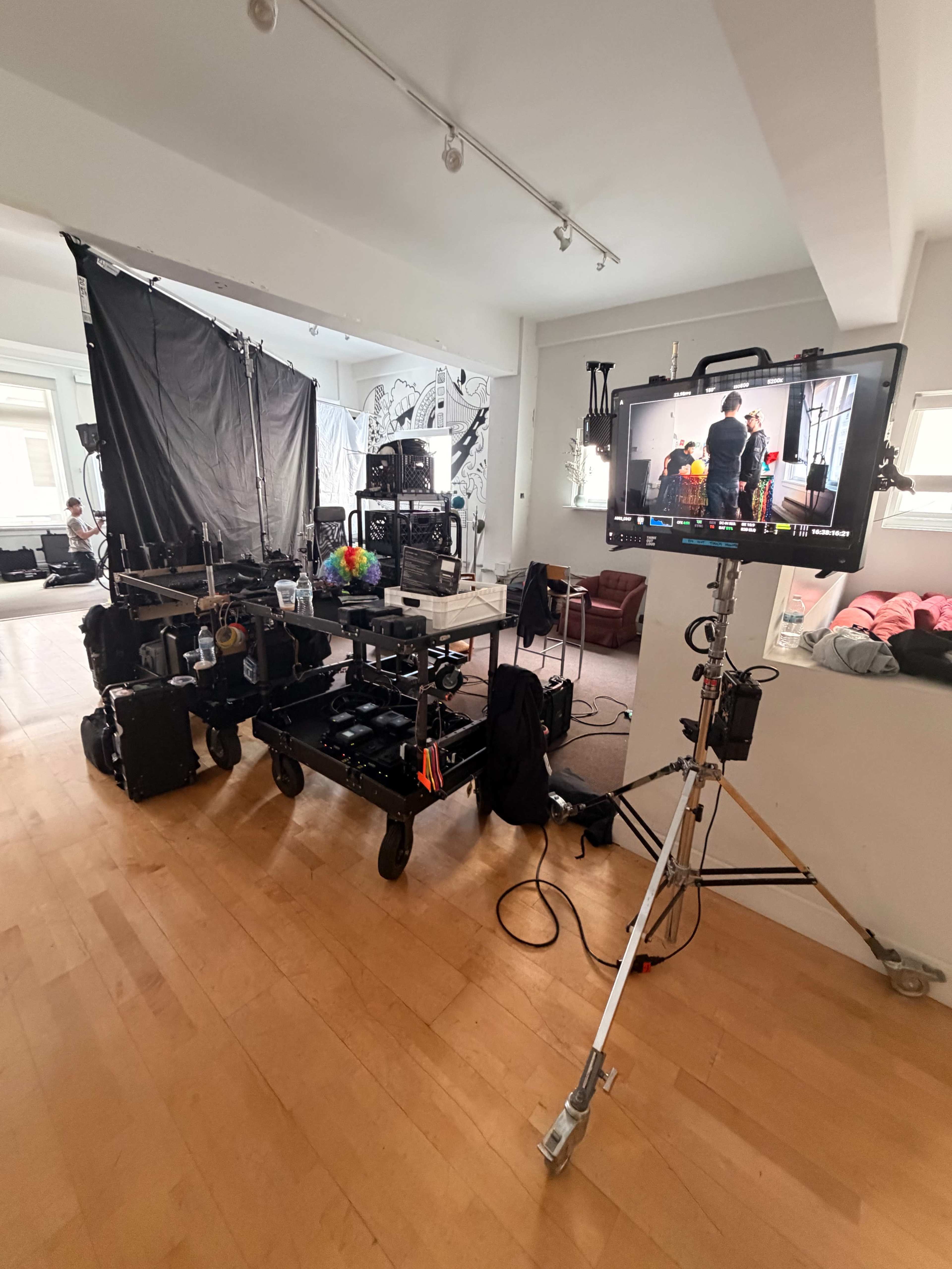 The image shows a film set with various equipment, including a monitor, lighting gear, and carts filled with gear, in a spacious room with light-colored walls.