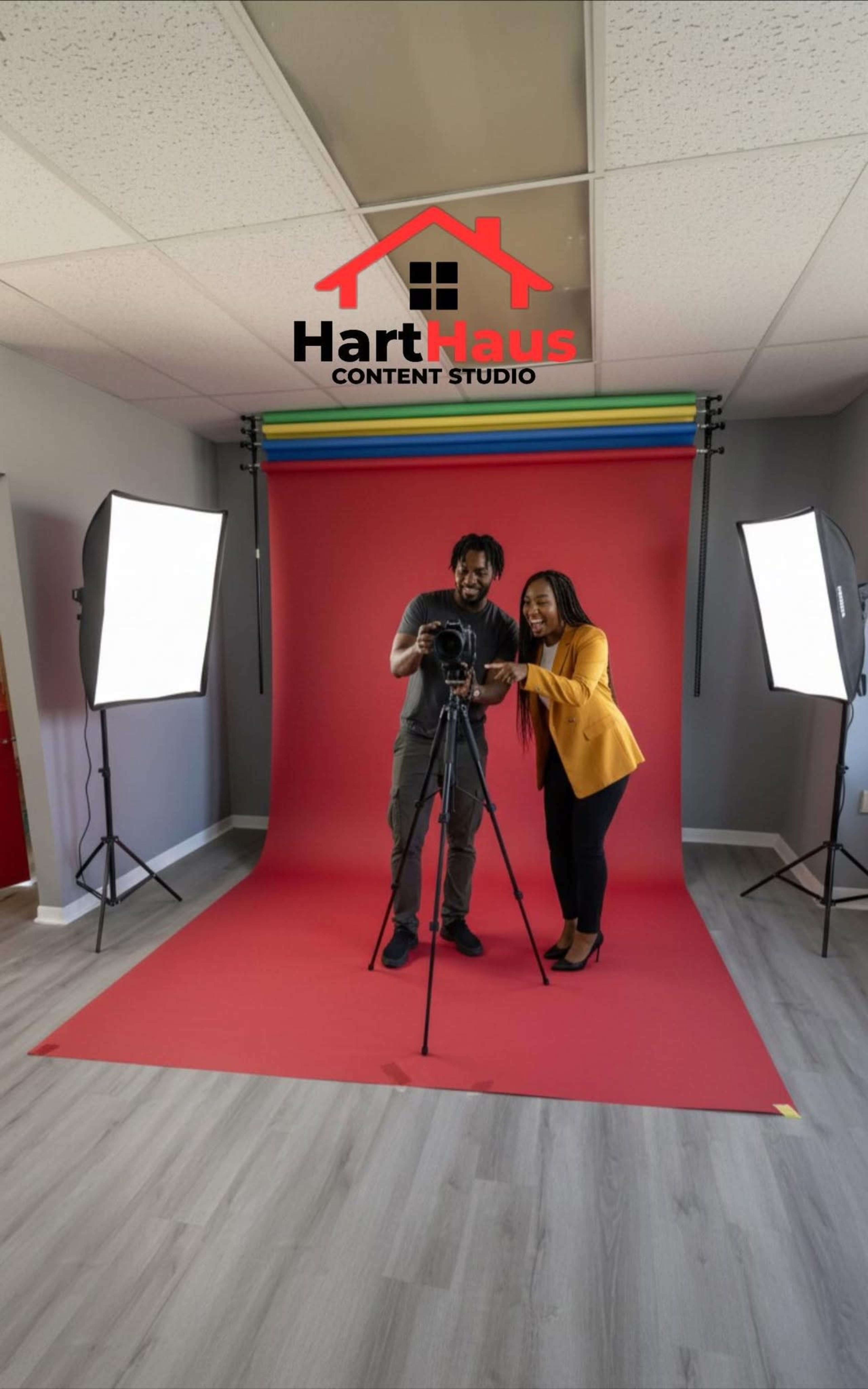 A photographer and a model pose together in a brightly lit studio with a red backdrop and professional lighting equipment.