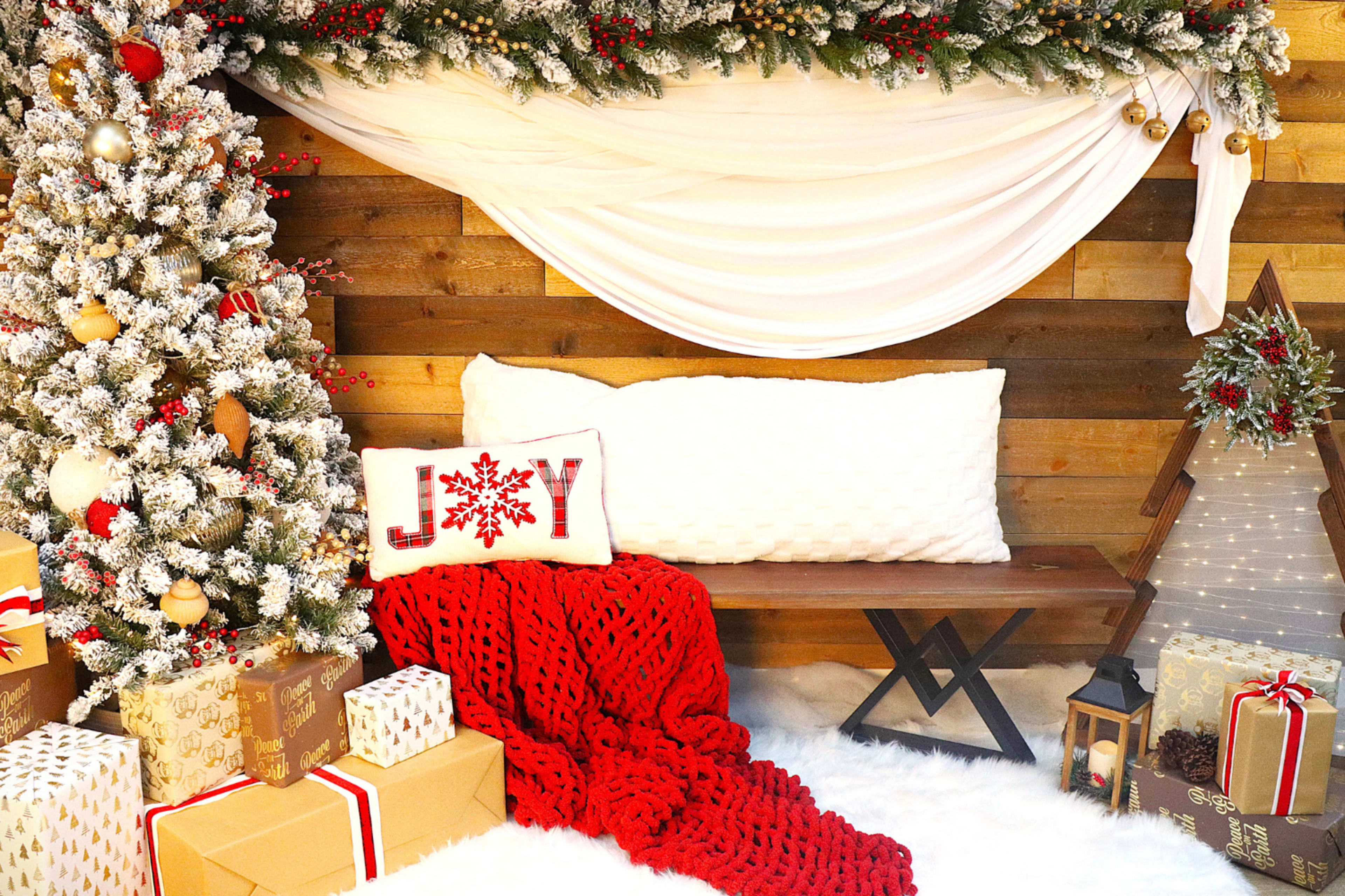 The image features a festive holiday scene with a decorated Christmas tree, a cozy red blanket draped over a wooden bench, and wrapped gifts arranged on a soft white rug.