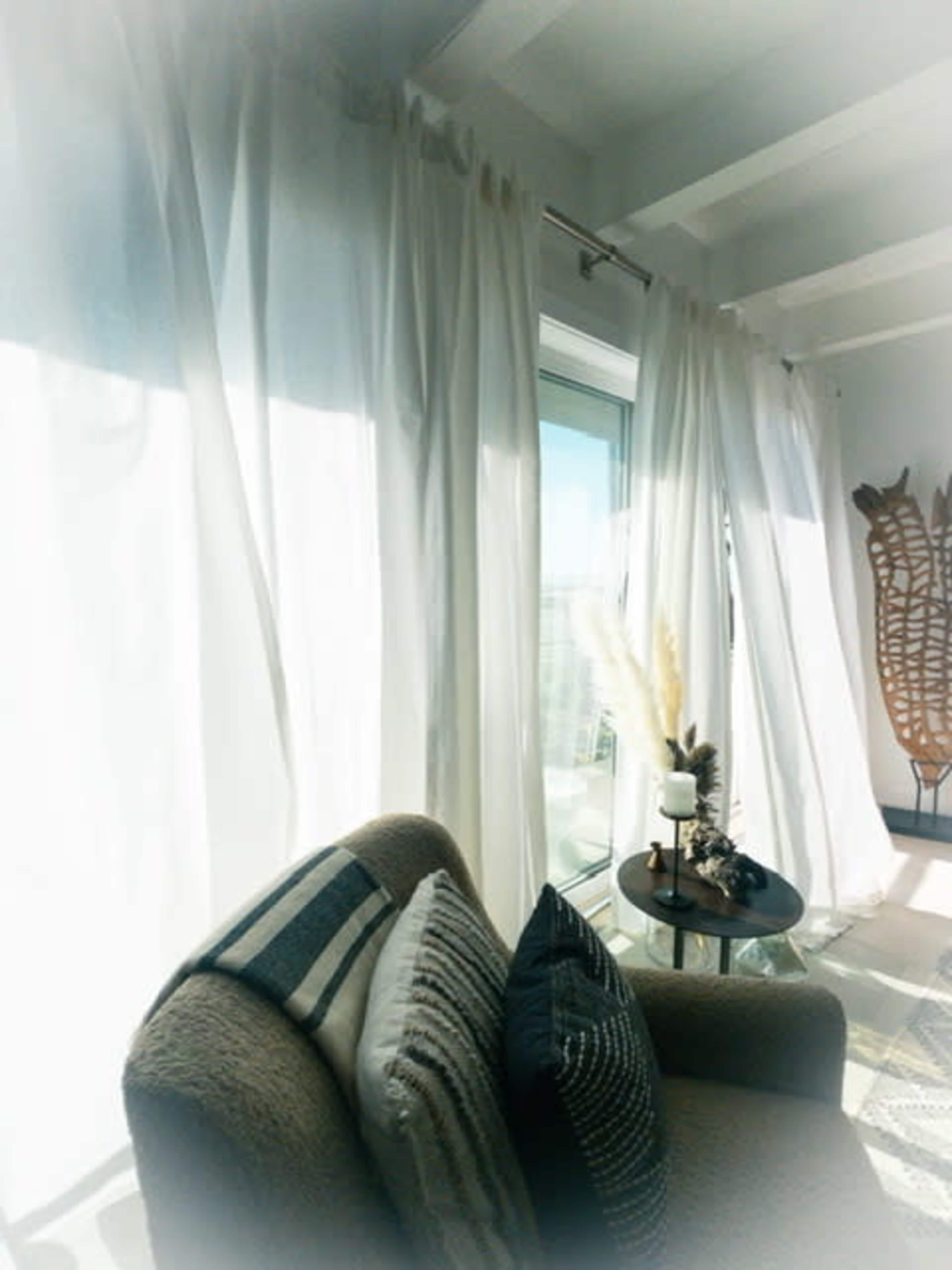 A cozy corner of a sunlit room features a soft armchair, decorative pillows, and sheer white curtains billowing near a small side table with decorative items.