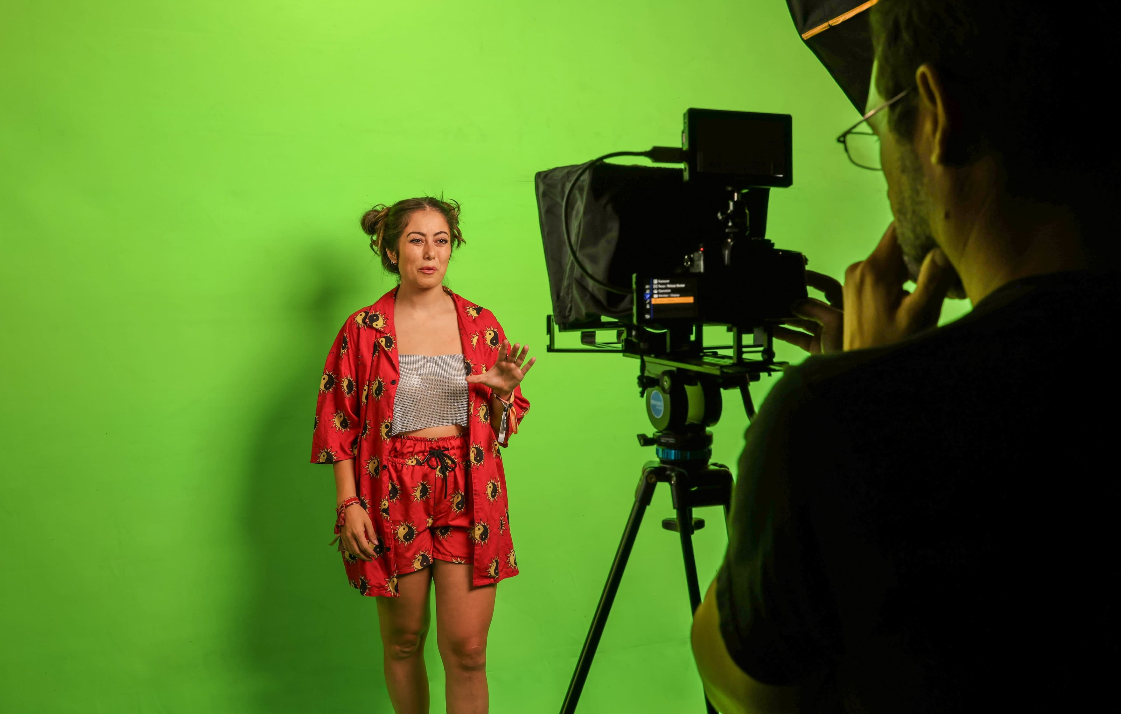 A woman stands in front of a green screen while a man operates a camera on a tripod.