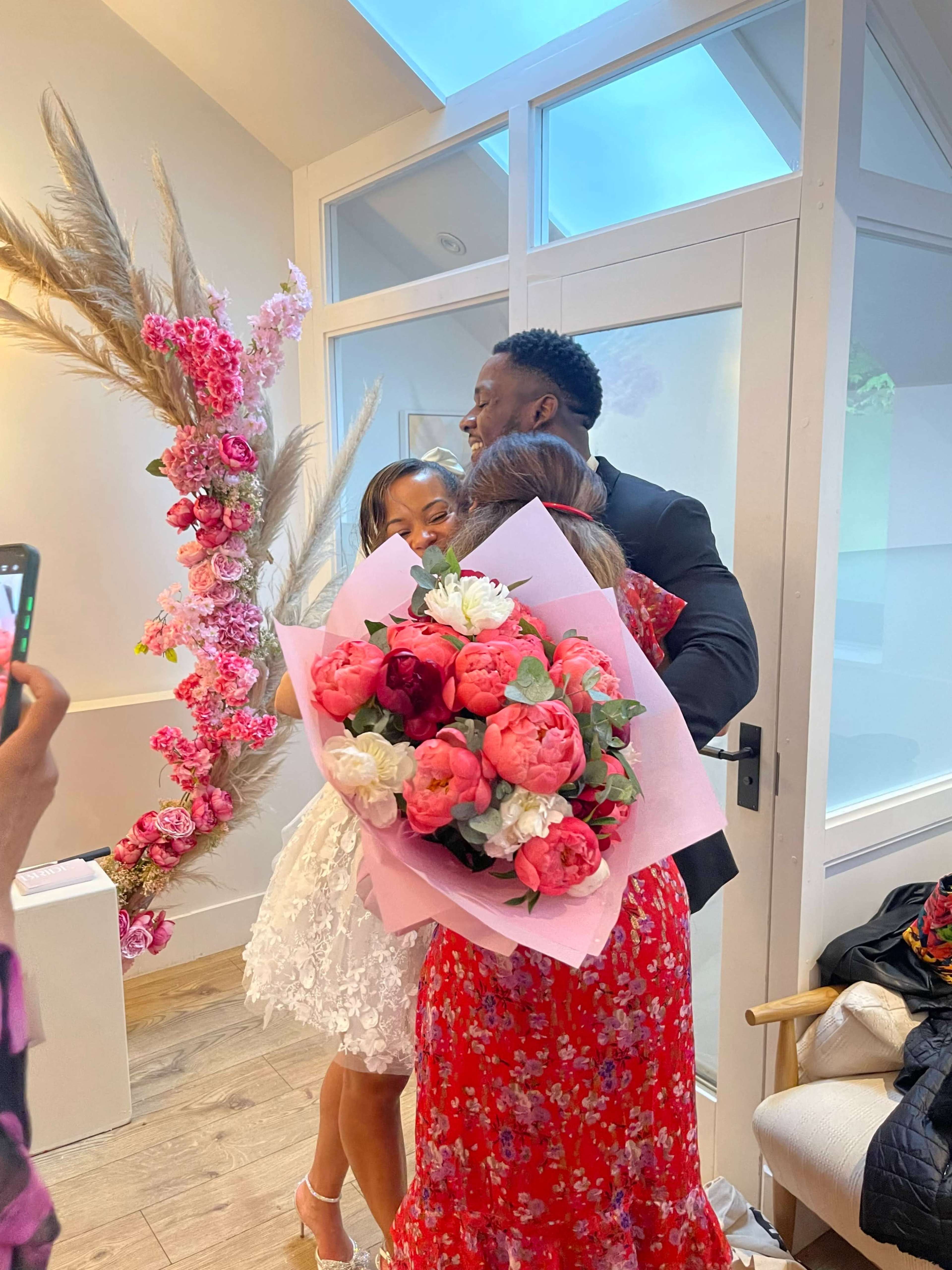 A woman in a white dress embraces a man in a suit while holding a large bouquet of pink flowers in a decorated room with floral arrangements.