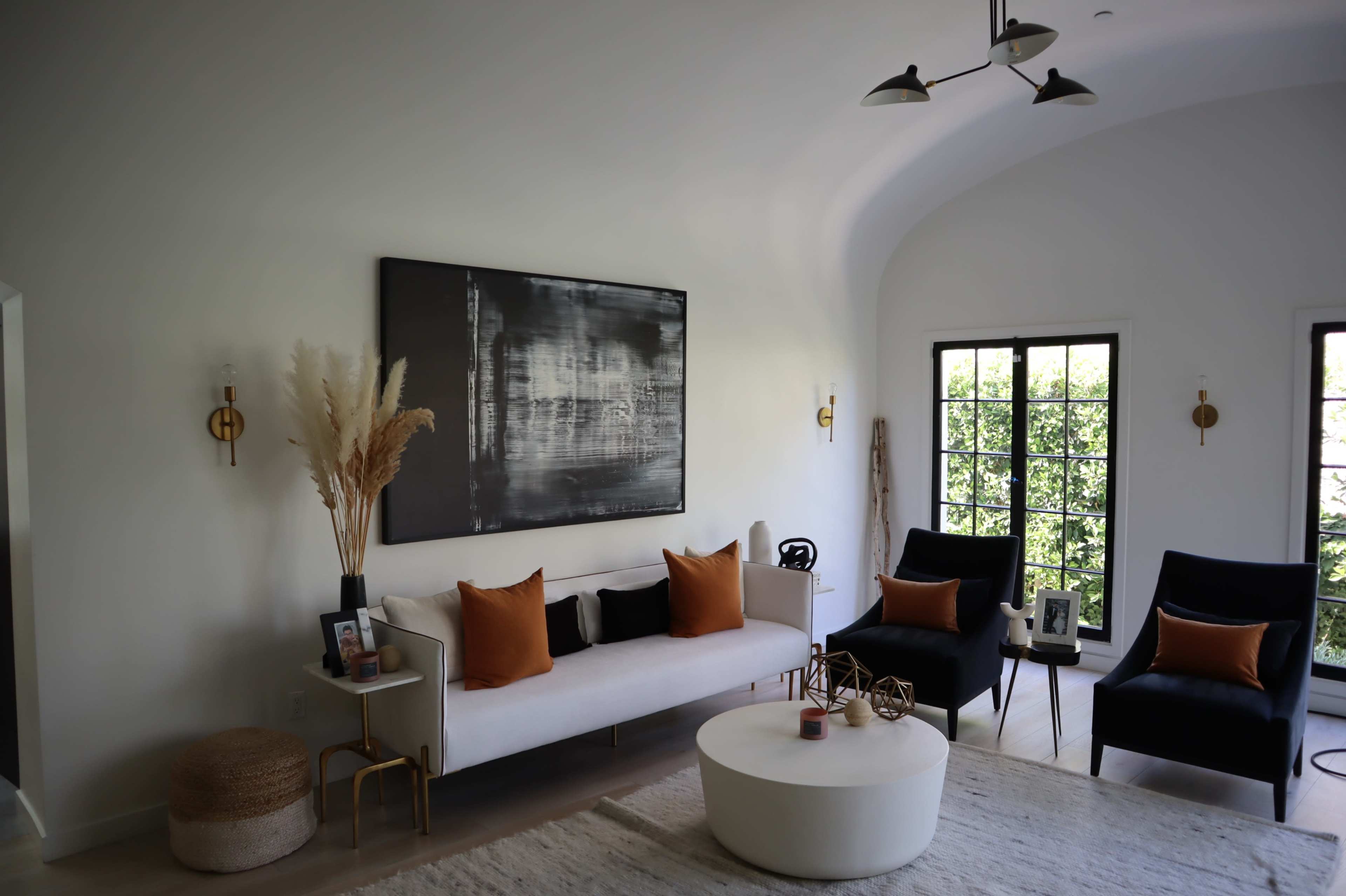 A bright living room features a white sofa with black and orange pillows, two dark chairs, a round coffee table, and framed windows overlooking greenery.