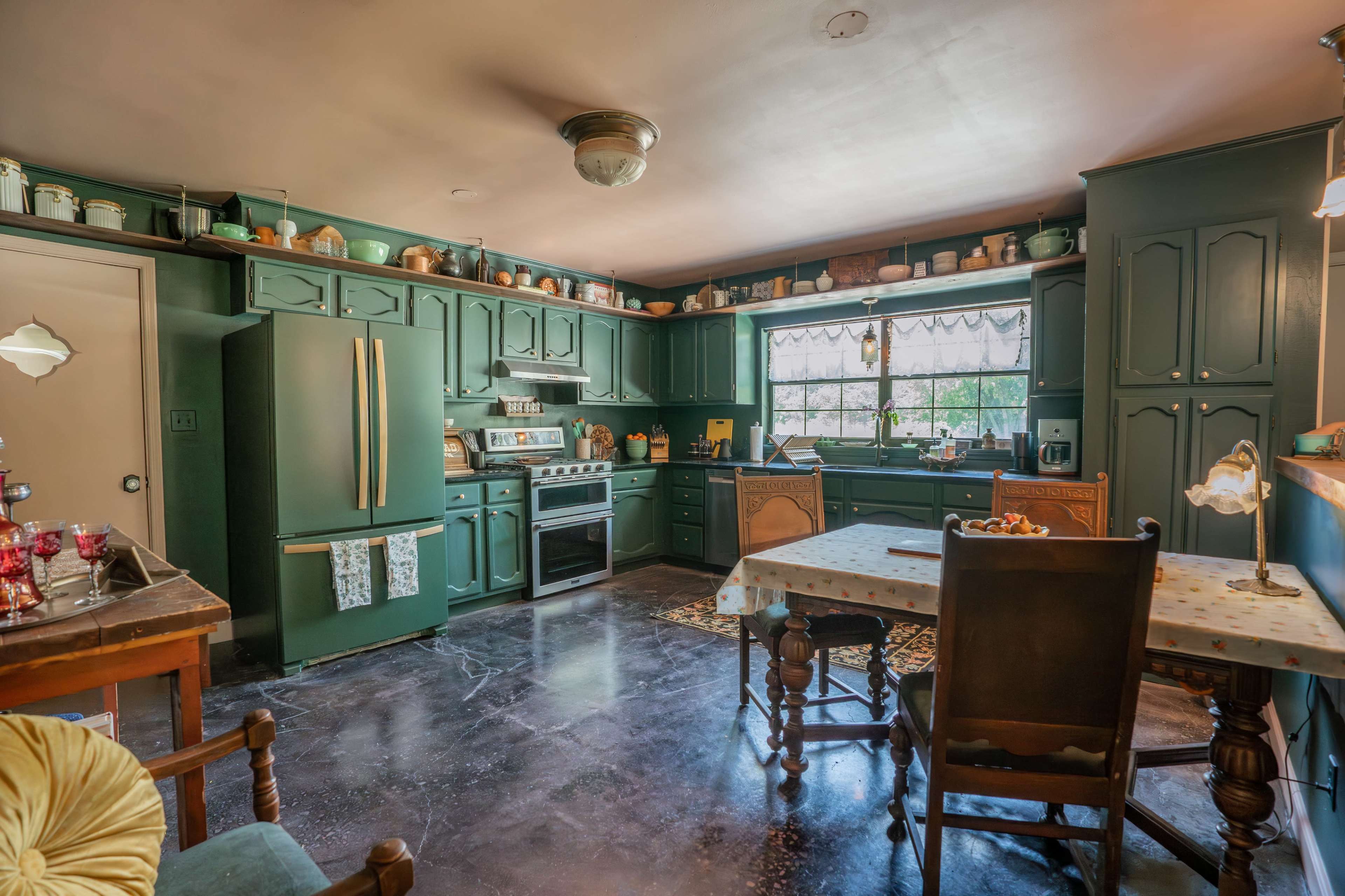 A dark green kitchen with wooden cabinets, a stove, a window, a dining table, and various kitchenware on the shelves.