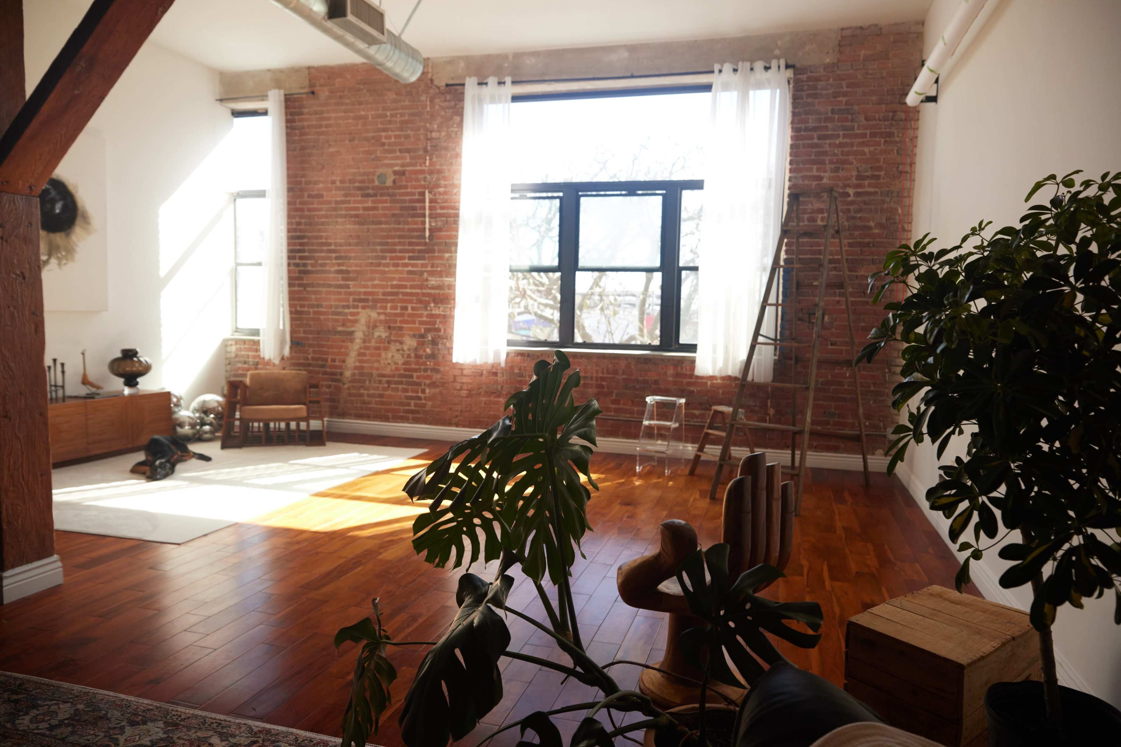 The image shows a spacious room with wooden floors, exposed brick walls, large windows with sheer curtains, and a ladder against the wall.