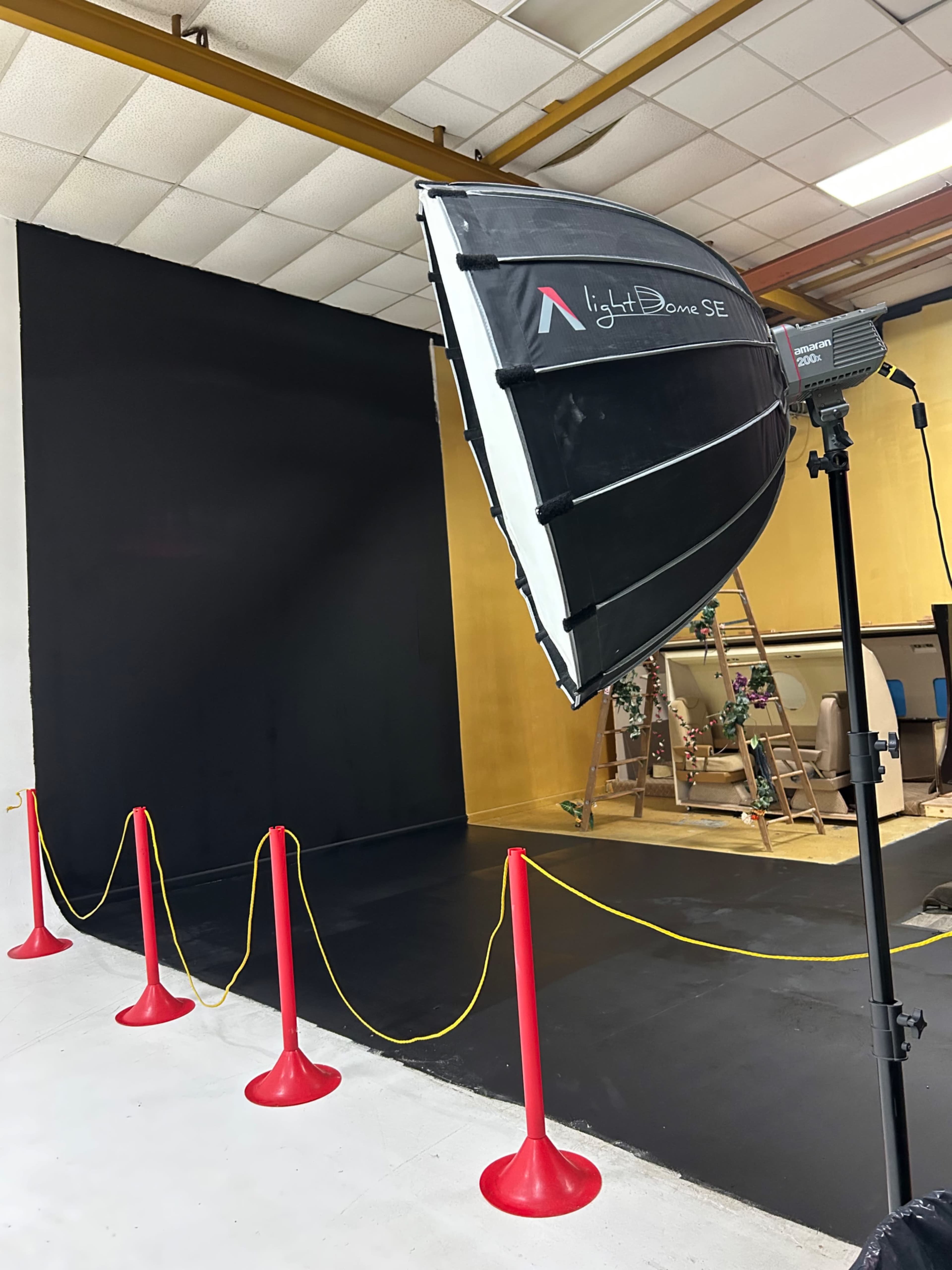 A photography studio setup features a large softbox lighting fixture, a black backdrop, and red barriers with yellow caution tape.