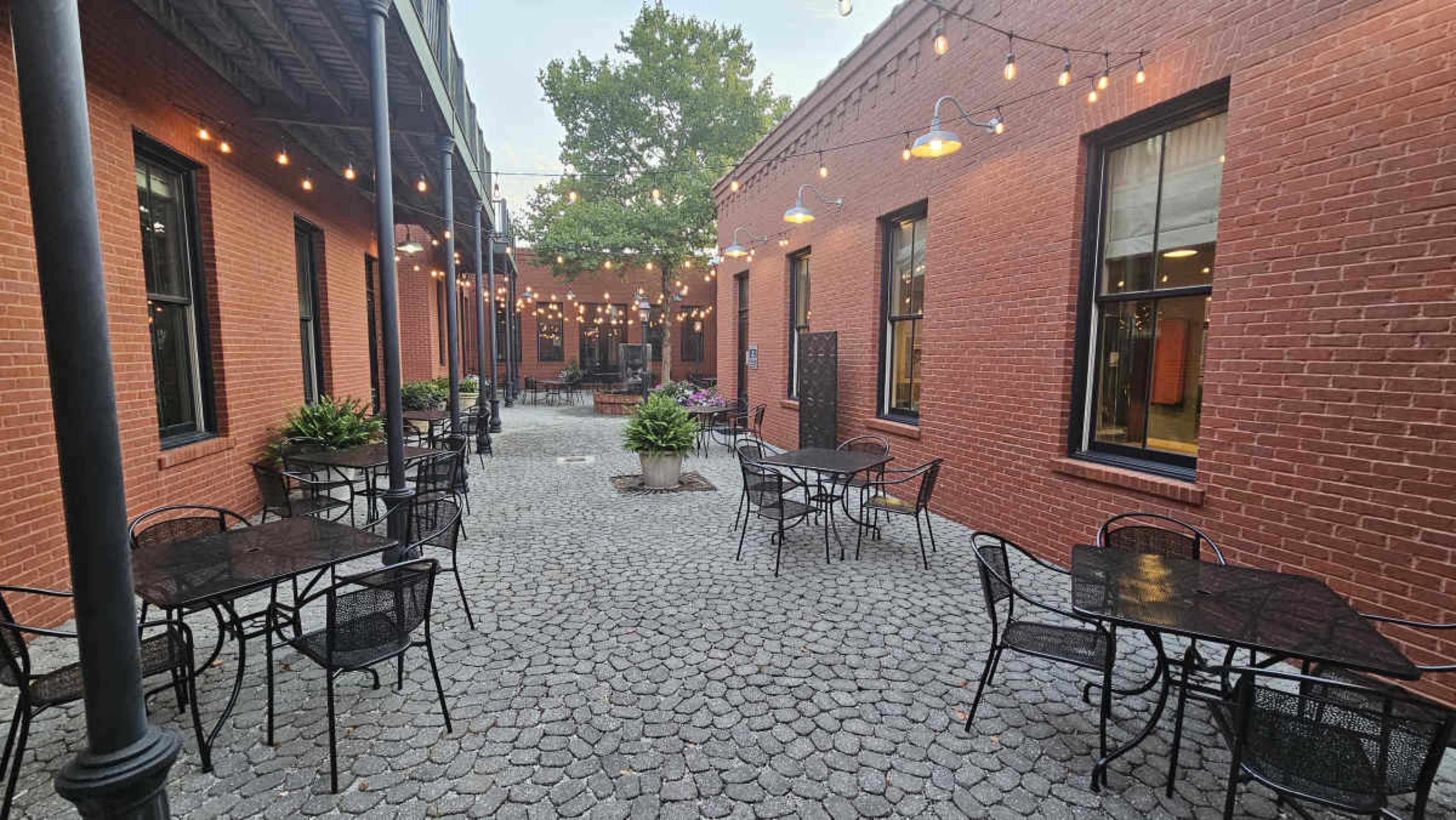 The image shows a cobblestone courtyard with several black metal tables and chairs surrounded by brick buildings, illuminated by overhead string lights.