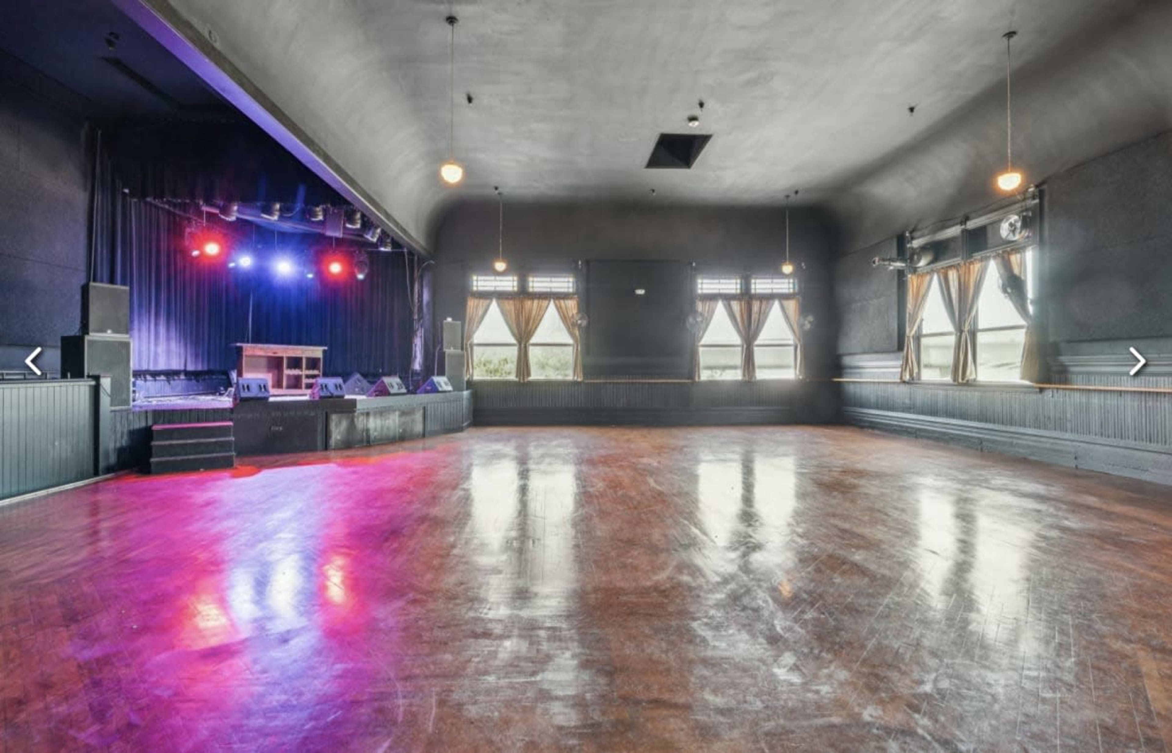 The image shows a spacious, empty performance venue with a stage at one end, large windows, and wooden flooring reflecting colored lights.