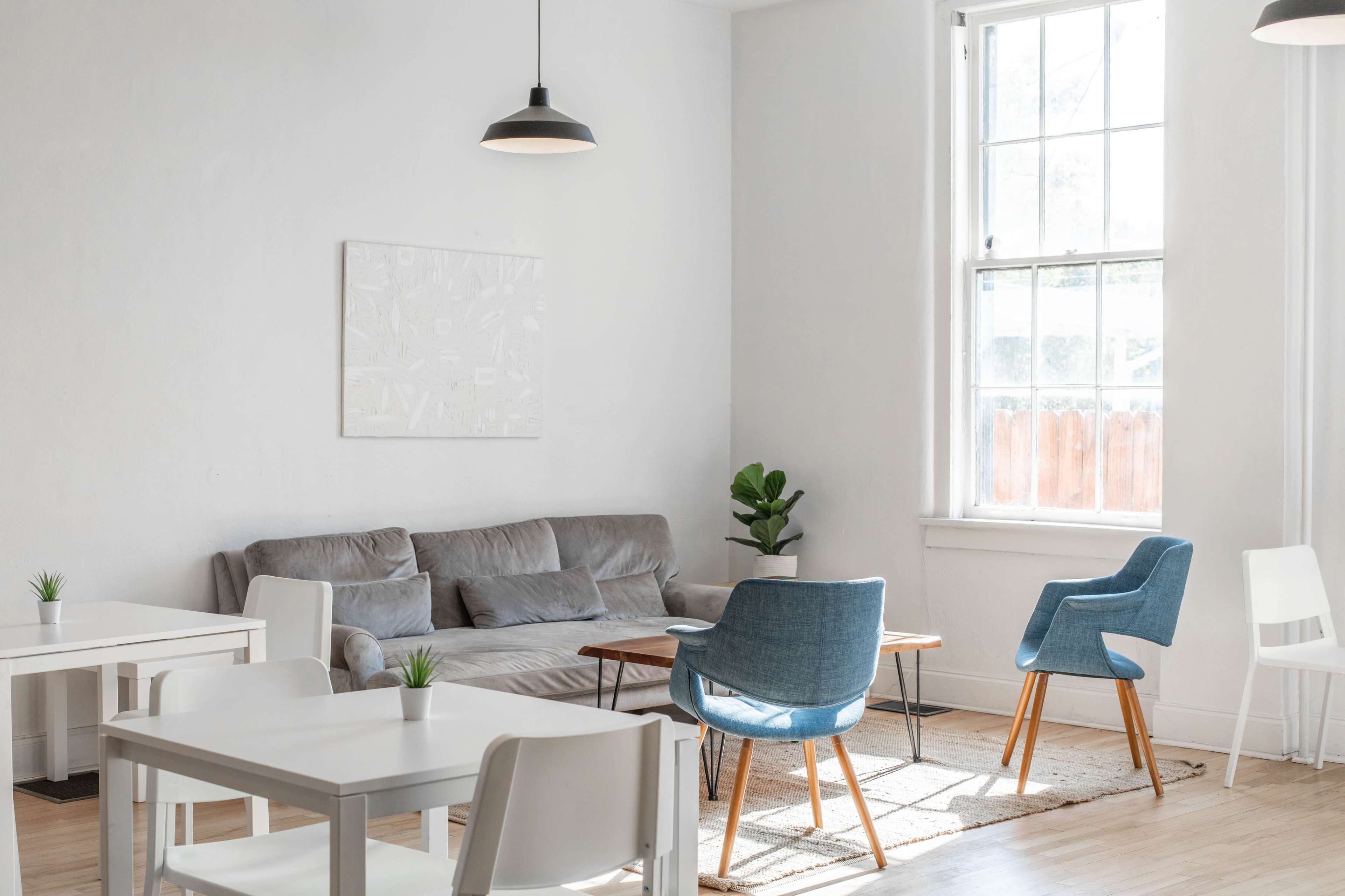 A minimalist living space with a gray sofa, a wooden coffee table, and blue chairs arranged around a light-colored area rug, illuminated by natural light from large windows.