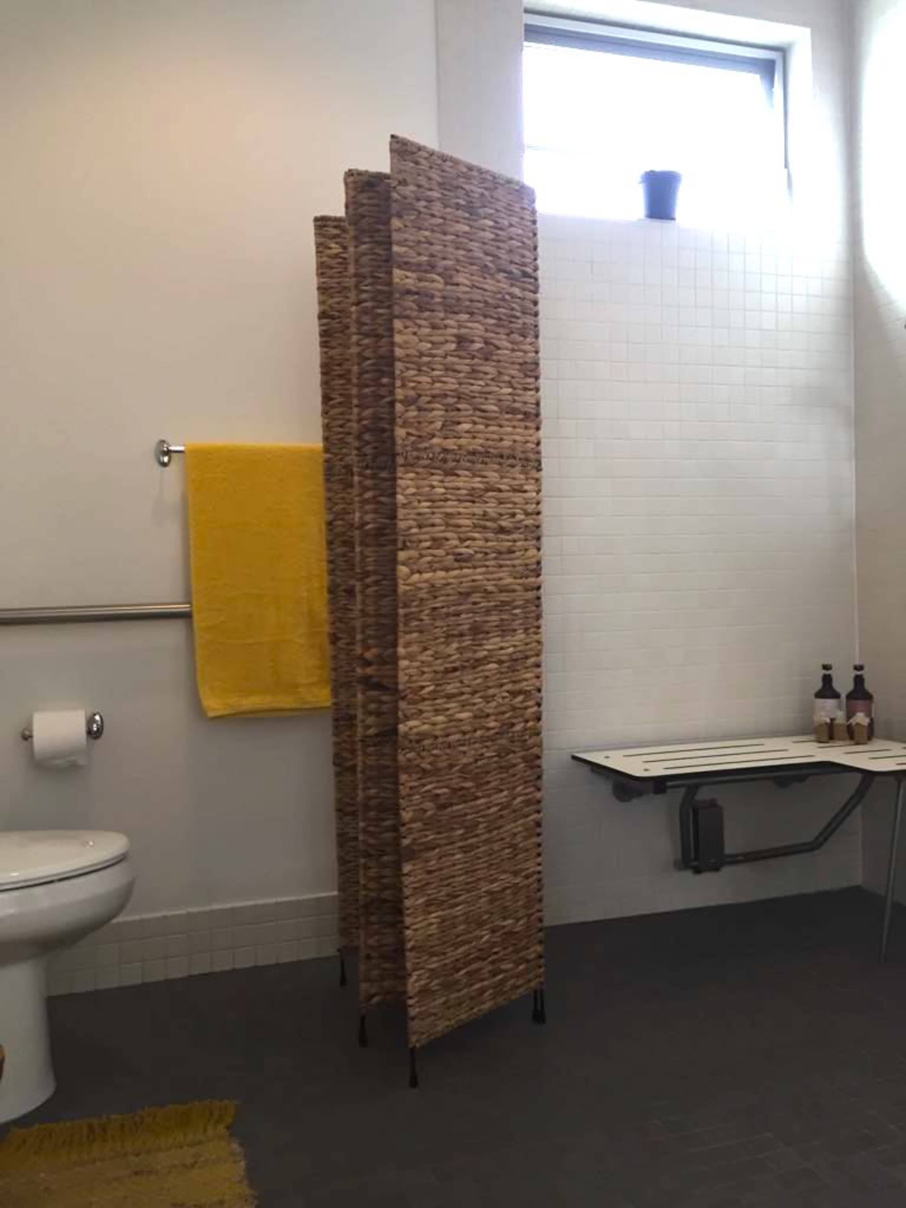 A bathroom with a wicker privacy screen, a yellow towel hanging on a rack, a toilet, and a folding table against the wall.