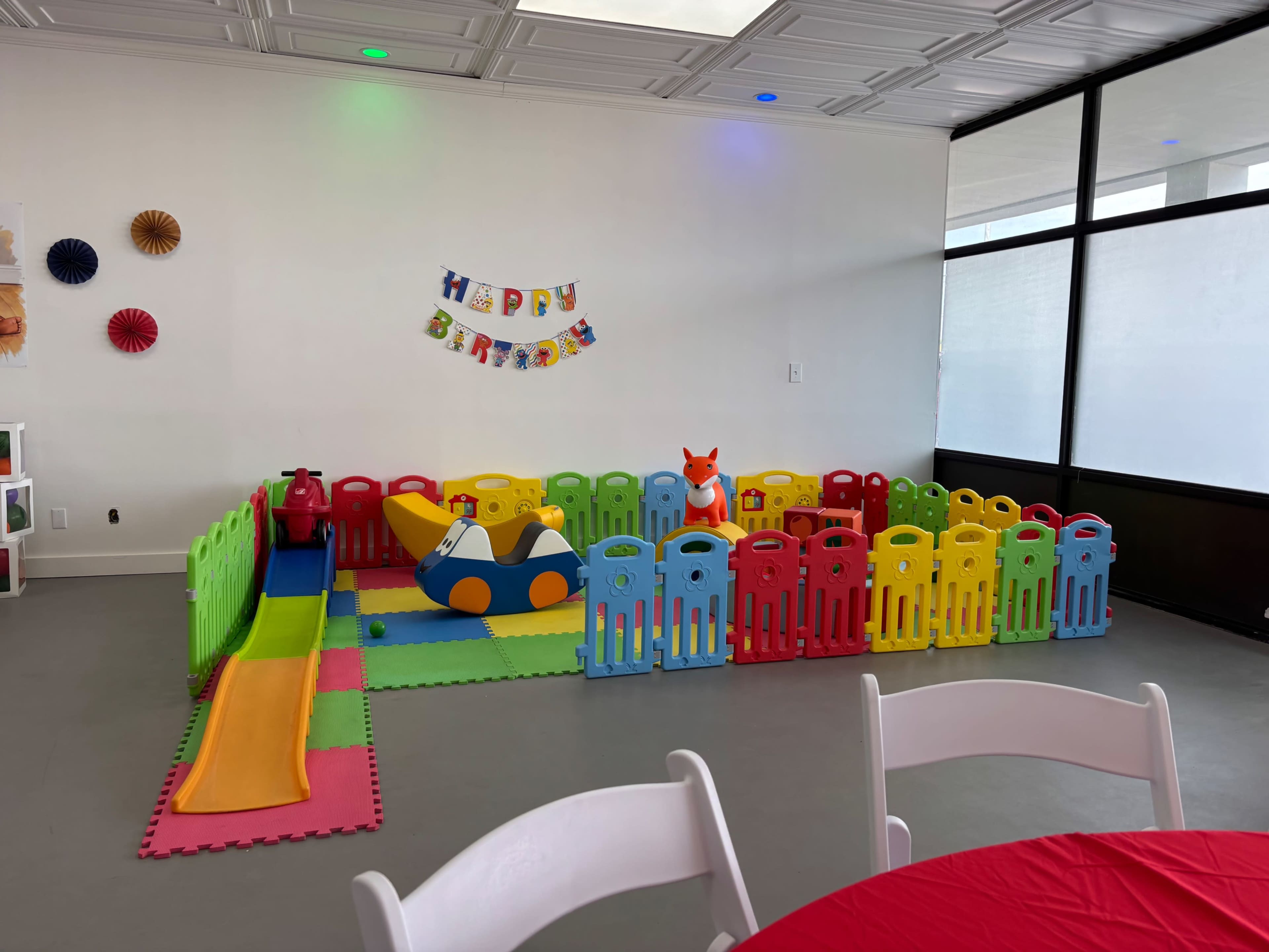 The image shows a colorful children's play area enclosed by plastic fences, featuring a slide, a toy car, and decorations for a birthday celebration.
