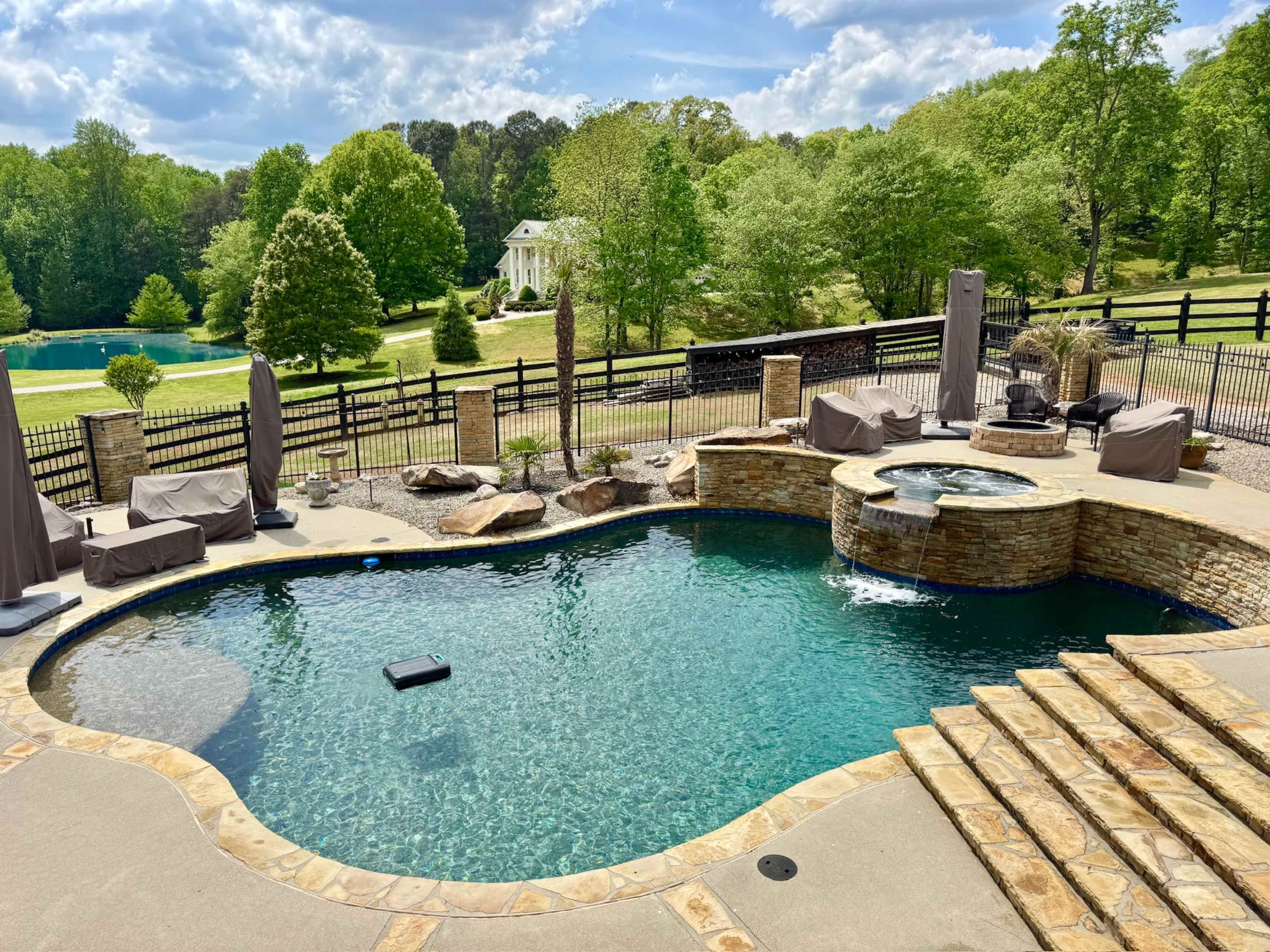 The image shows a spacious outdoor swimming pool surrounded by stone steps, lounge chairs, and greenery, under a partly cloudy sky.