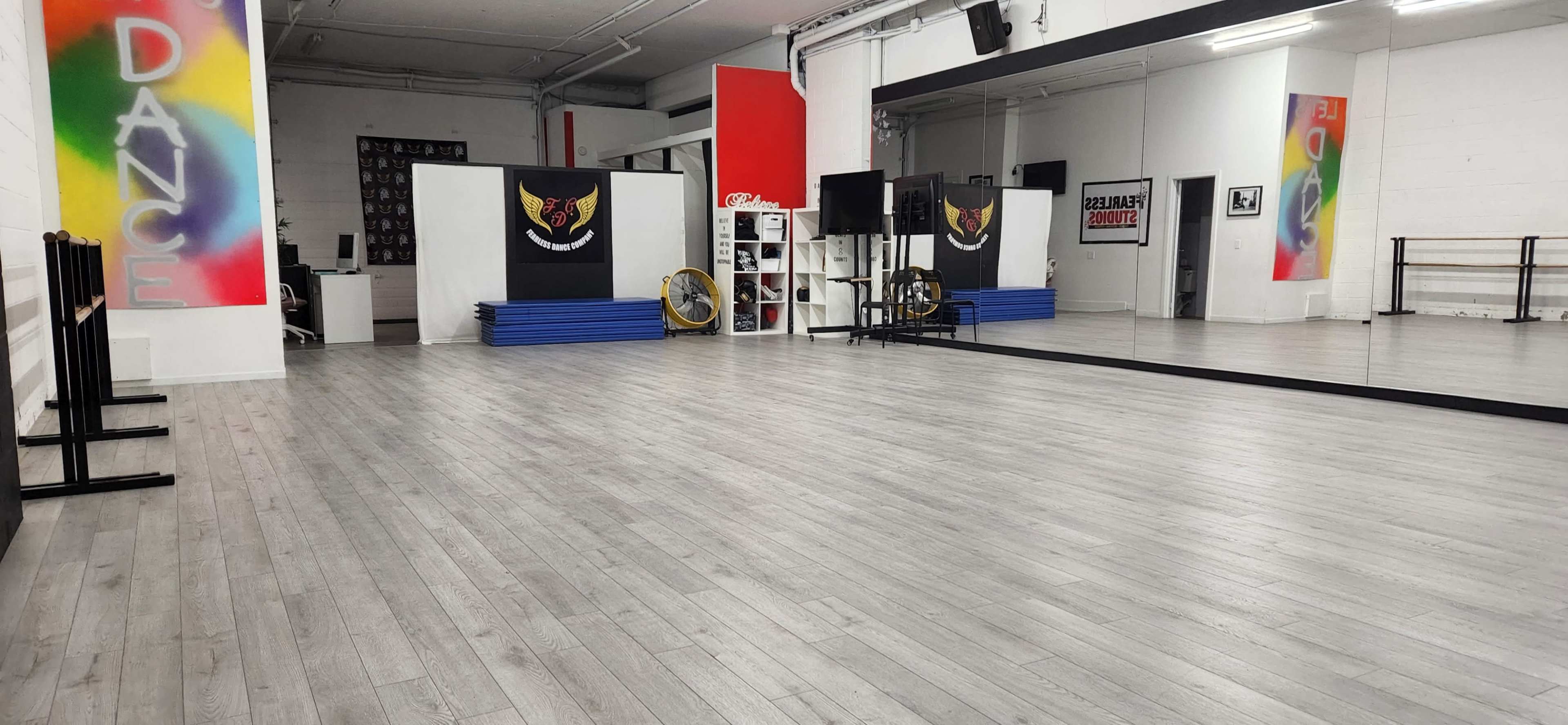 The image shows an empty dance studio with gray wooden floors, mirrors, and colorful wall murals.