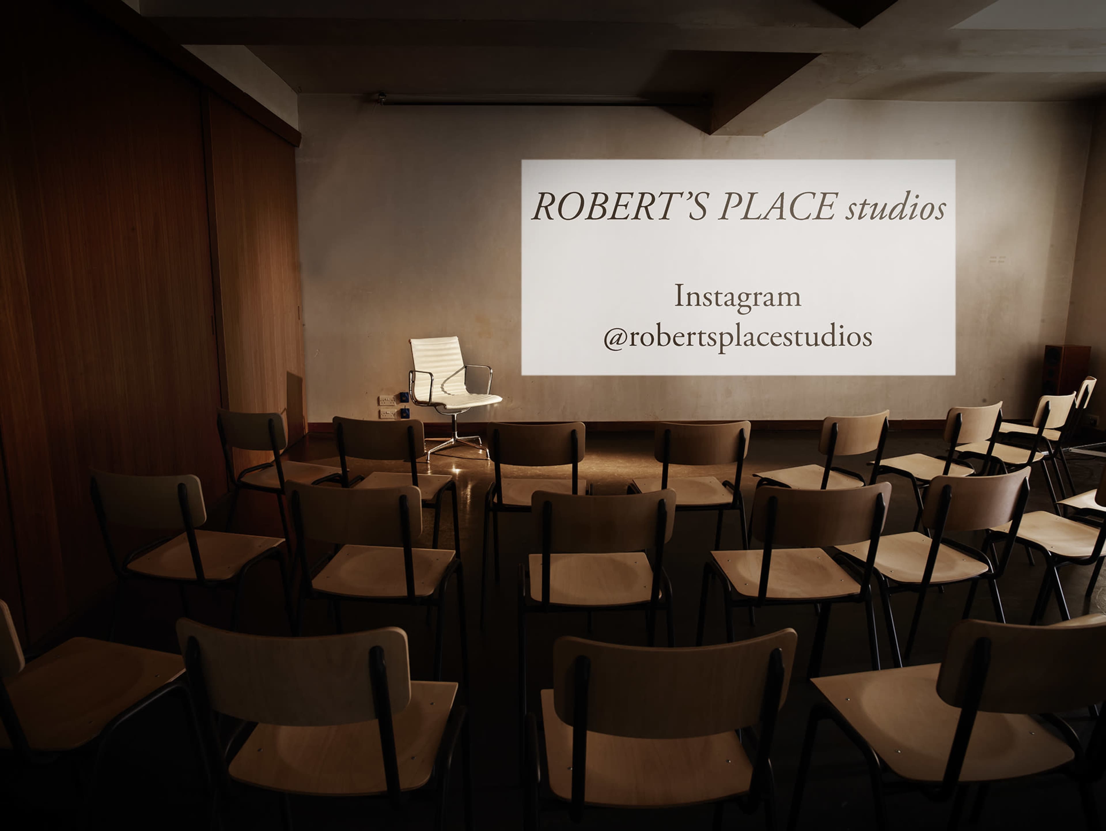 An empty conference room features a white projection screen with "ROBERT'S PLACE studios" displayed and a single chair in front of a row of wooden chairs.