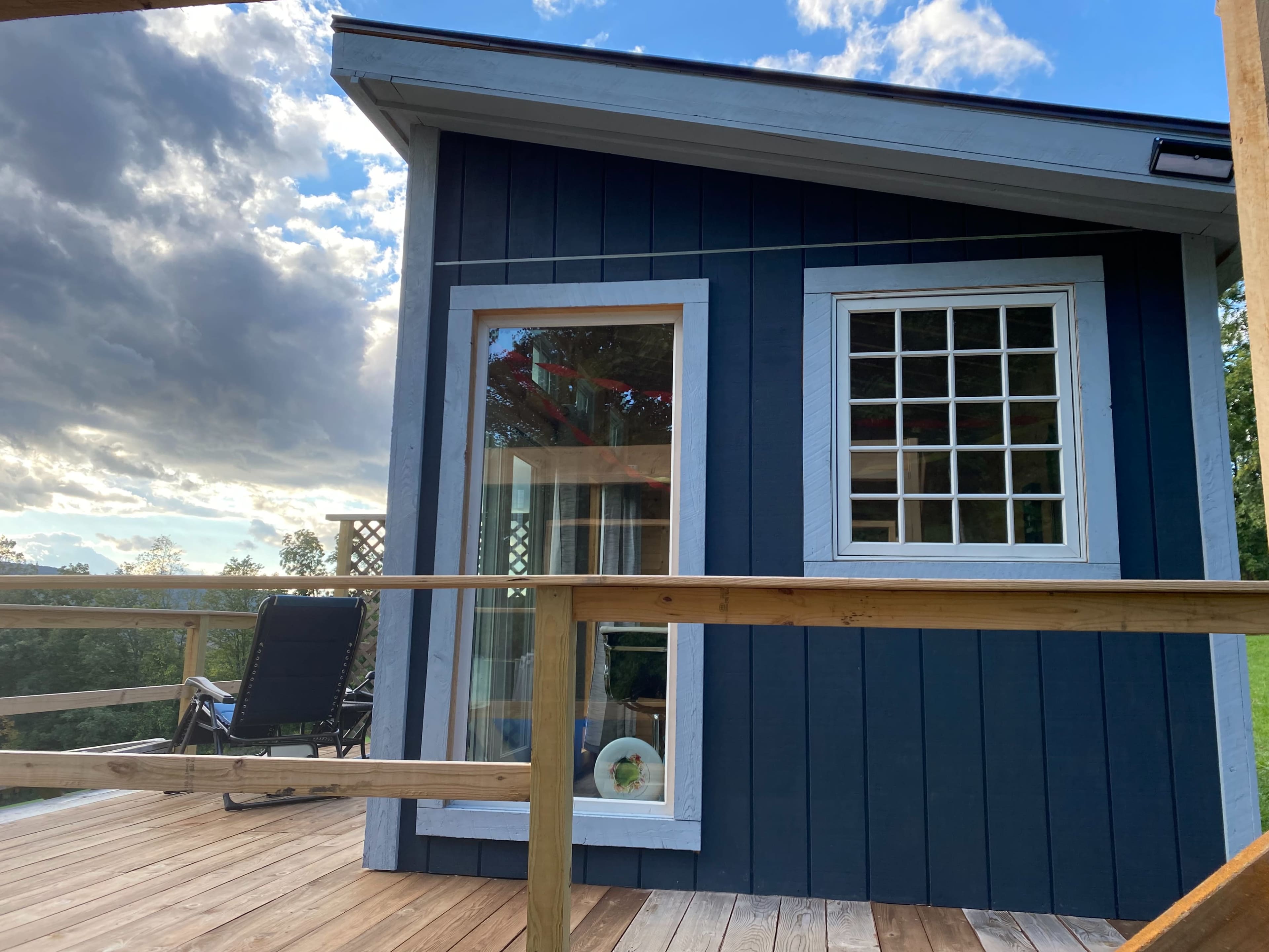 A dark blue wooden cabin with a large window and a deck overlooking a grassy area, under a partly cloudy sky.