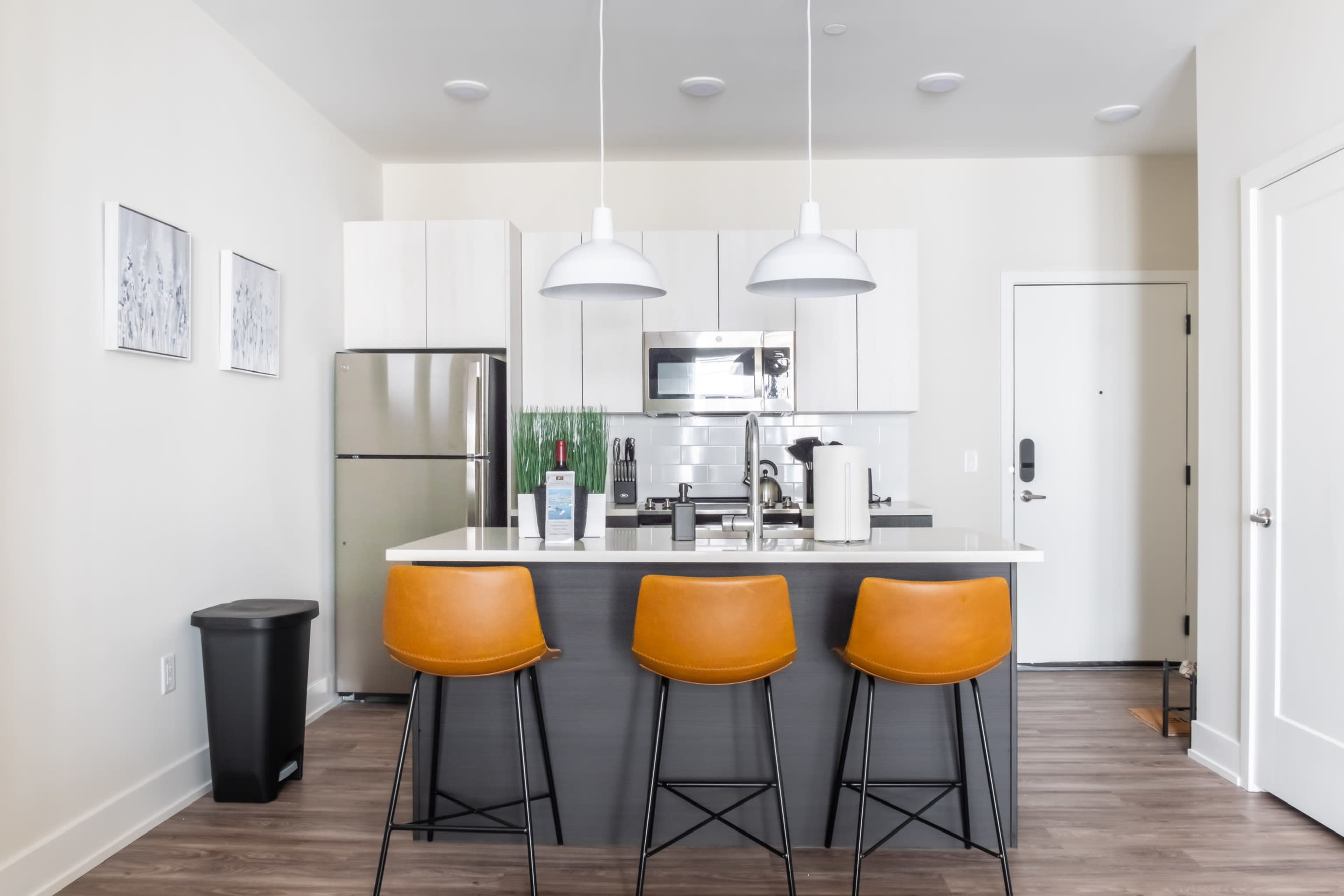 A modern kitchen features a central island with three brown bar stools, stainless steel appliances, and a door leading to a hallway.