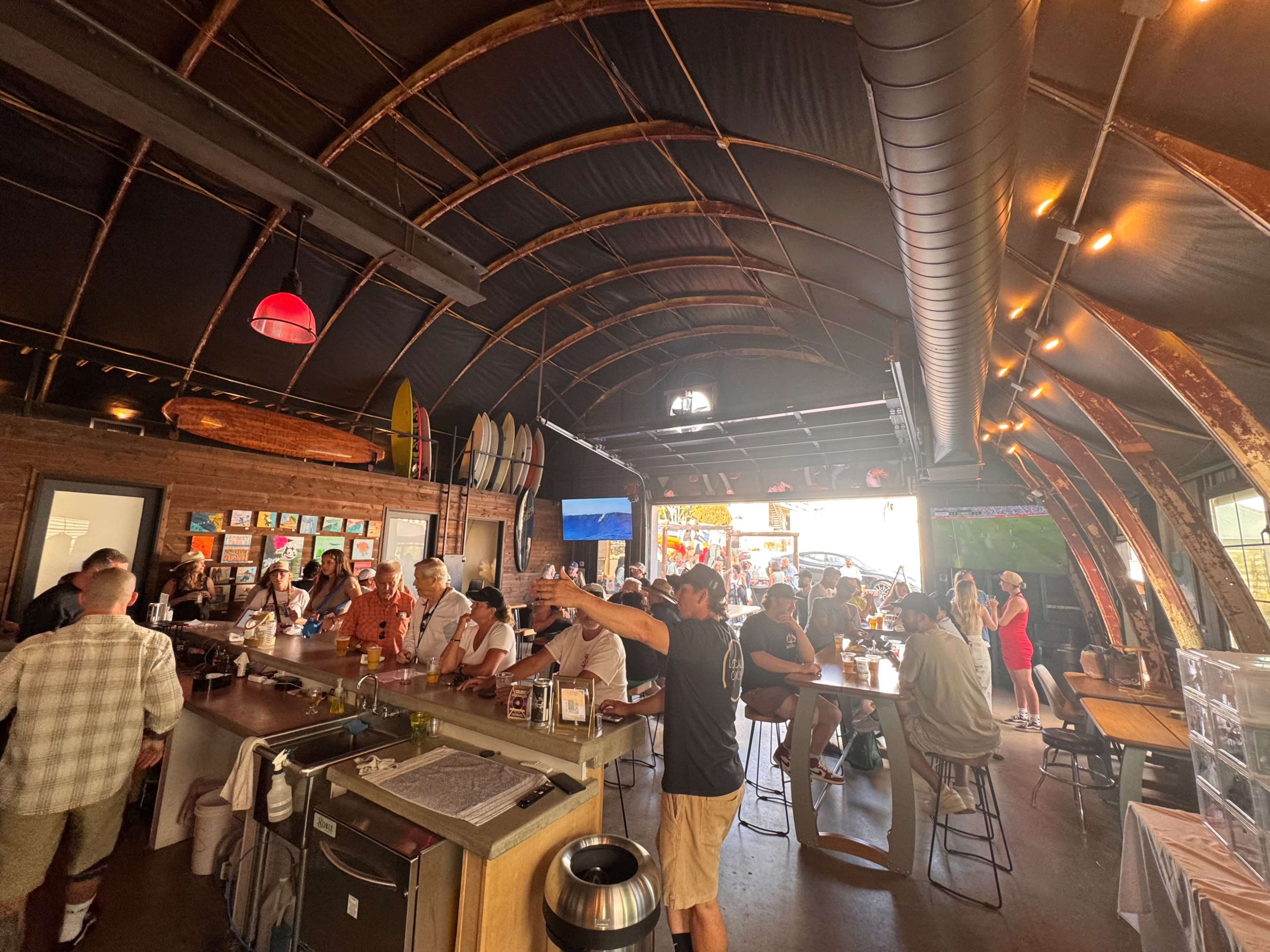 A bustling bar scene is depicted, with patrons socializing at high-top tables and the bar counter, surrounded by surfboards and an open space.
