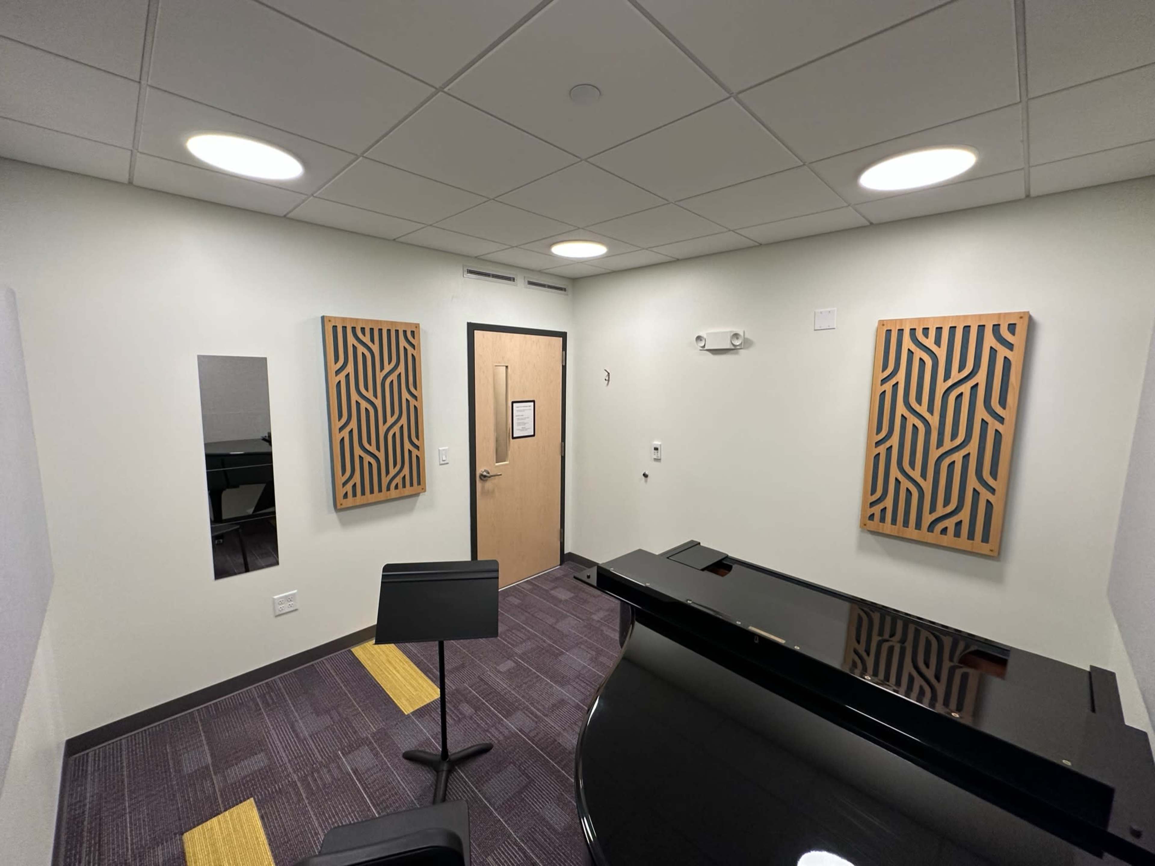 A small, well-lit music practice room featuring a grand piano, sound-absorbing wall panels, and a music stand.