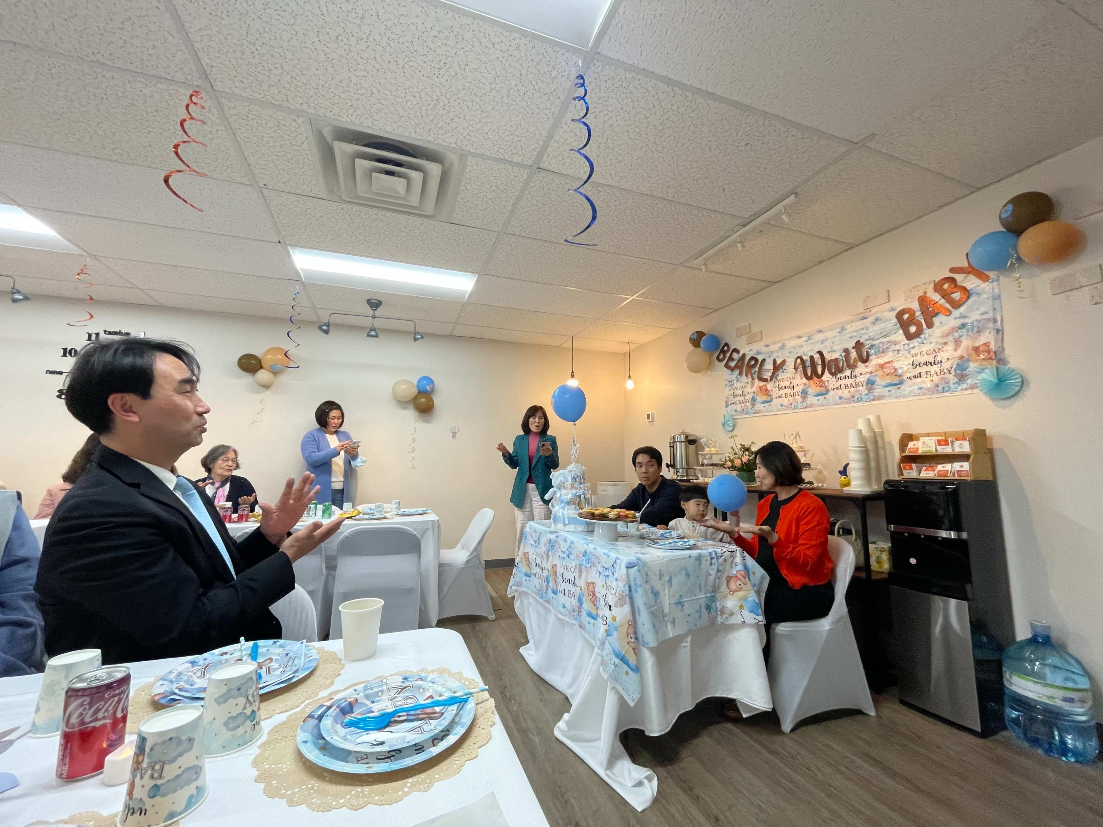 A small gathering is taking place in a decorated room with balloons and a "Baby" banner, where several people are seated and participating in celebration activities.