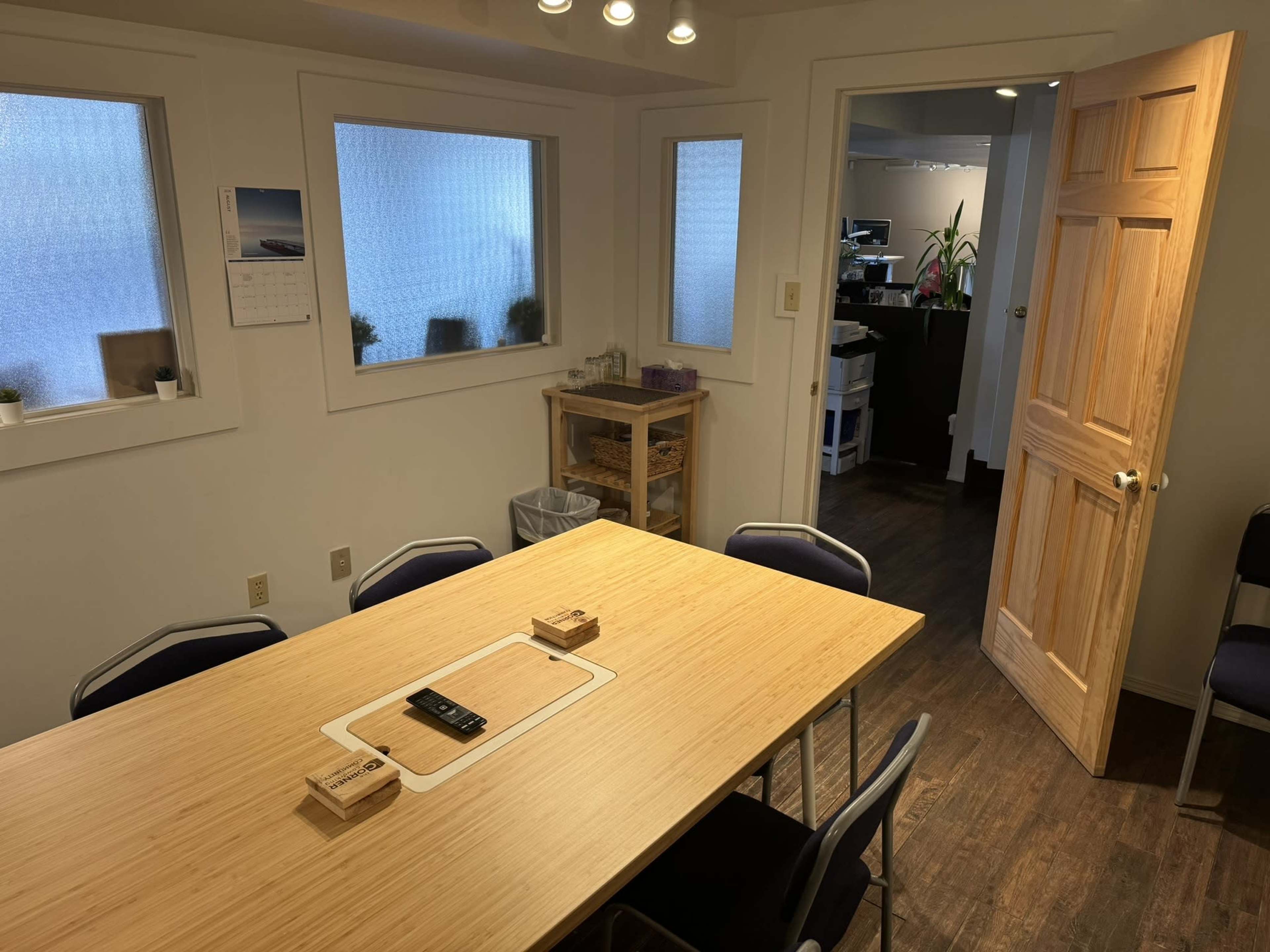 A simple meeting room with a wooden table surrounded by chairs, a calendar on the wall, and a door leading to another room.