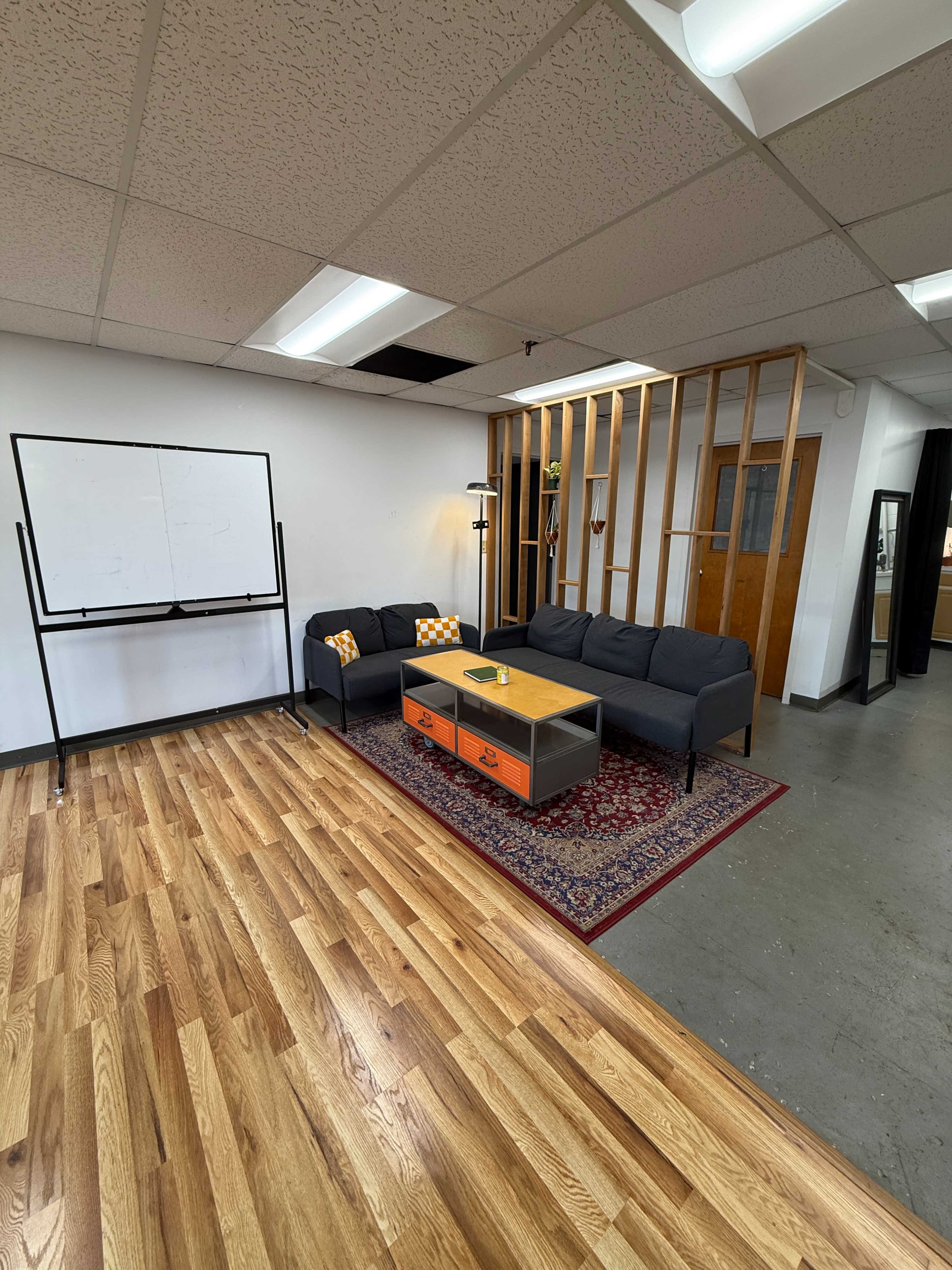 The image shows a modern office lounge area with a dark sofa, an orange coffee table, a large whiteboard, and wooden flooring.