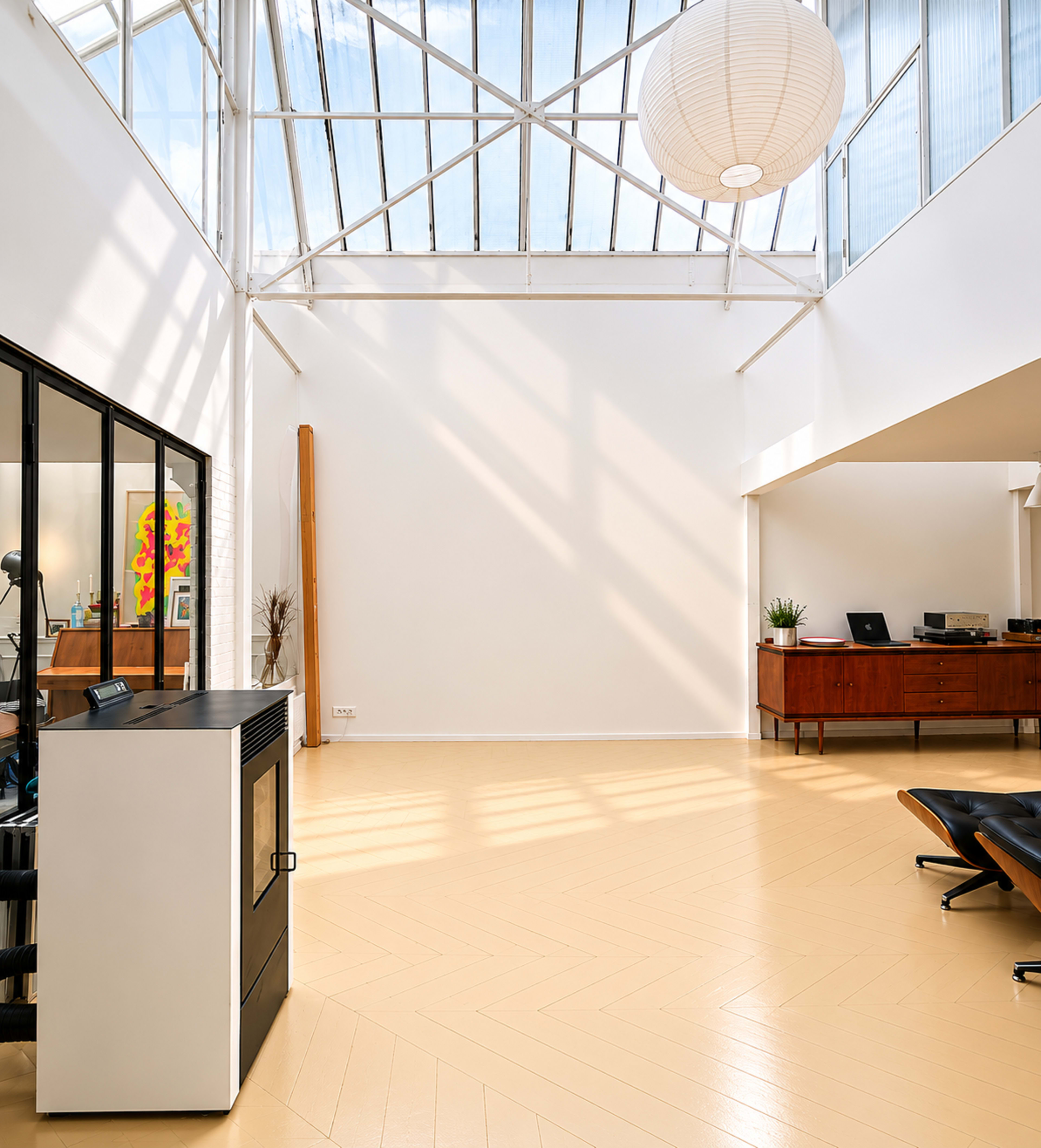 The image shows a bright, minimalist interior space with large windows, a wooden floor, and modern furniture including a black chair and a sideboard.