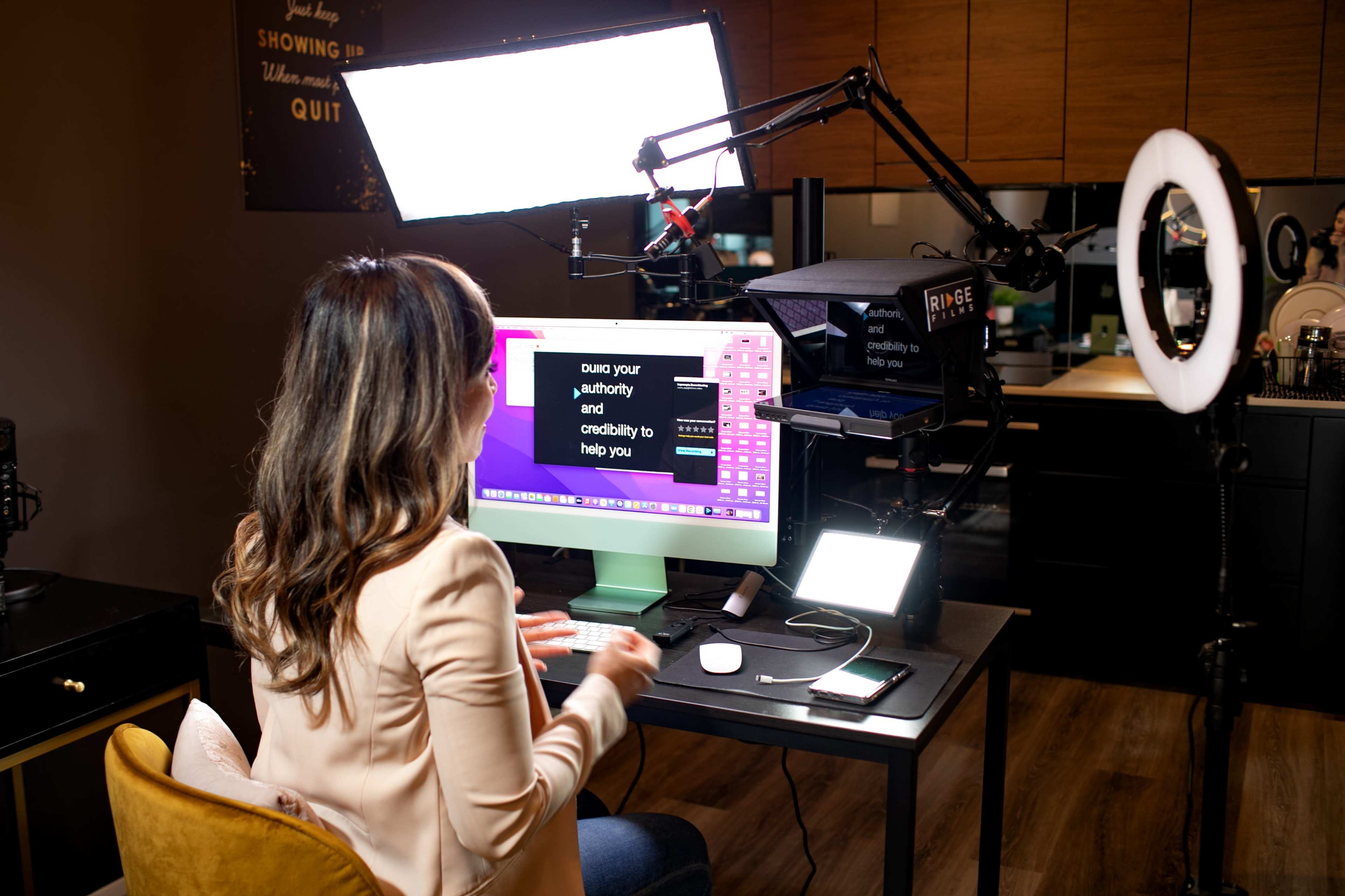 A person is sitting at a desk using a computer, with overhead lighting and filming equipment set up in a modern indoor space.