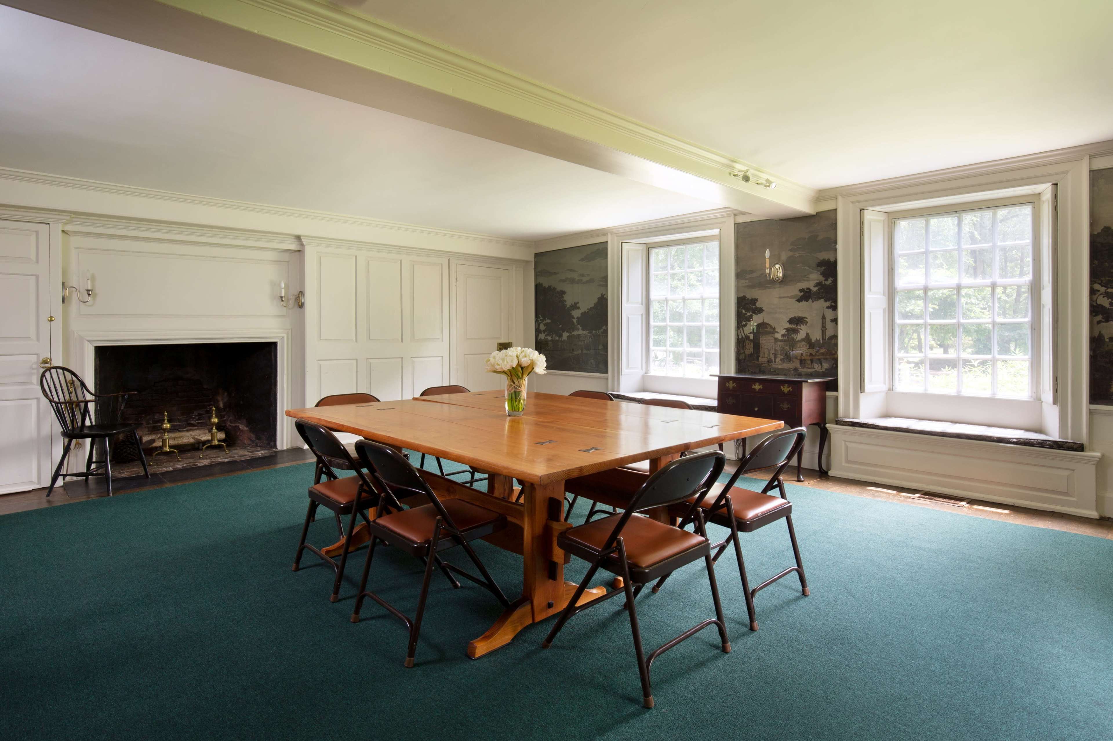A wooden table surrounded by black chairs is positioned in a room with a fireplace and large windows featuring a mural on the walls.