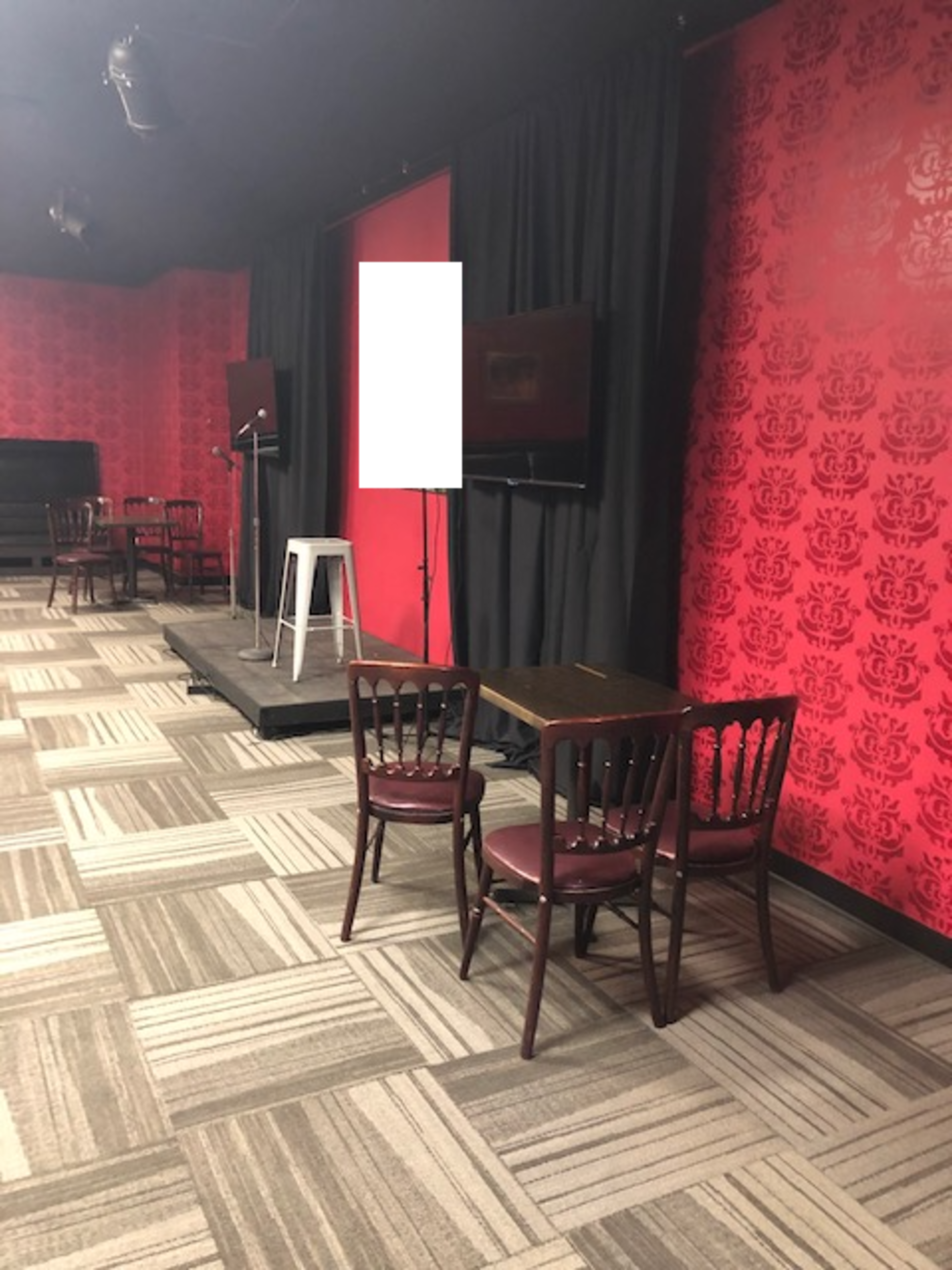 A small stage area with two microphones, a blank screen, and a setup of tables and chairs against a red patterned wall.