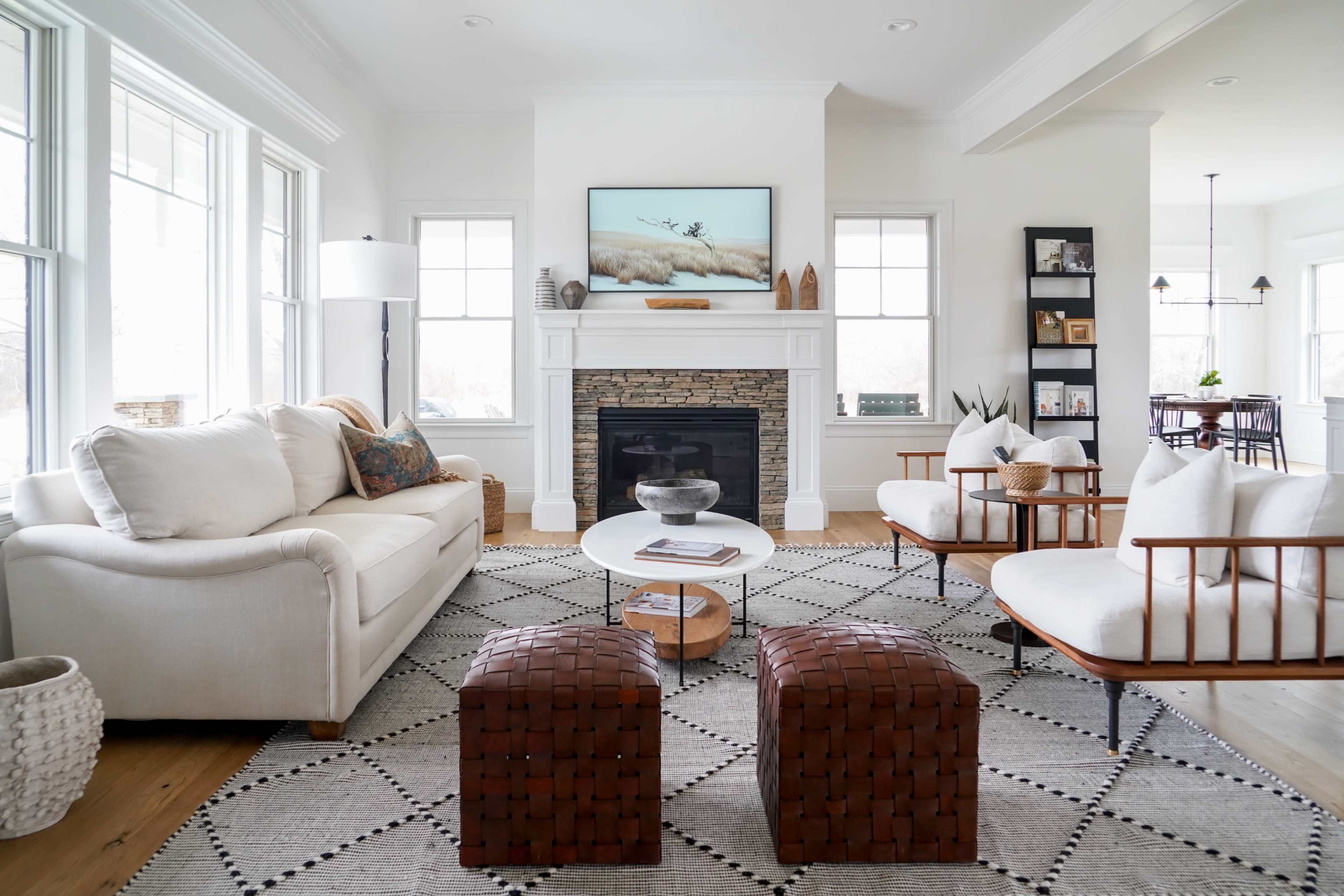 A bright living room with a white sofa, two wooden chairs, a round coffee table, and a fireplace under a television.