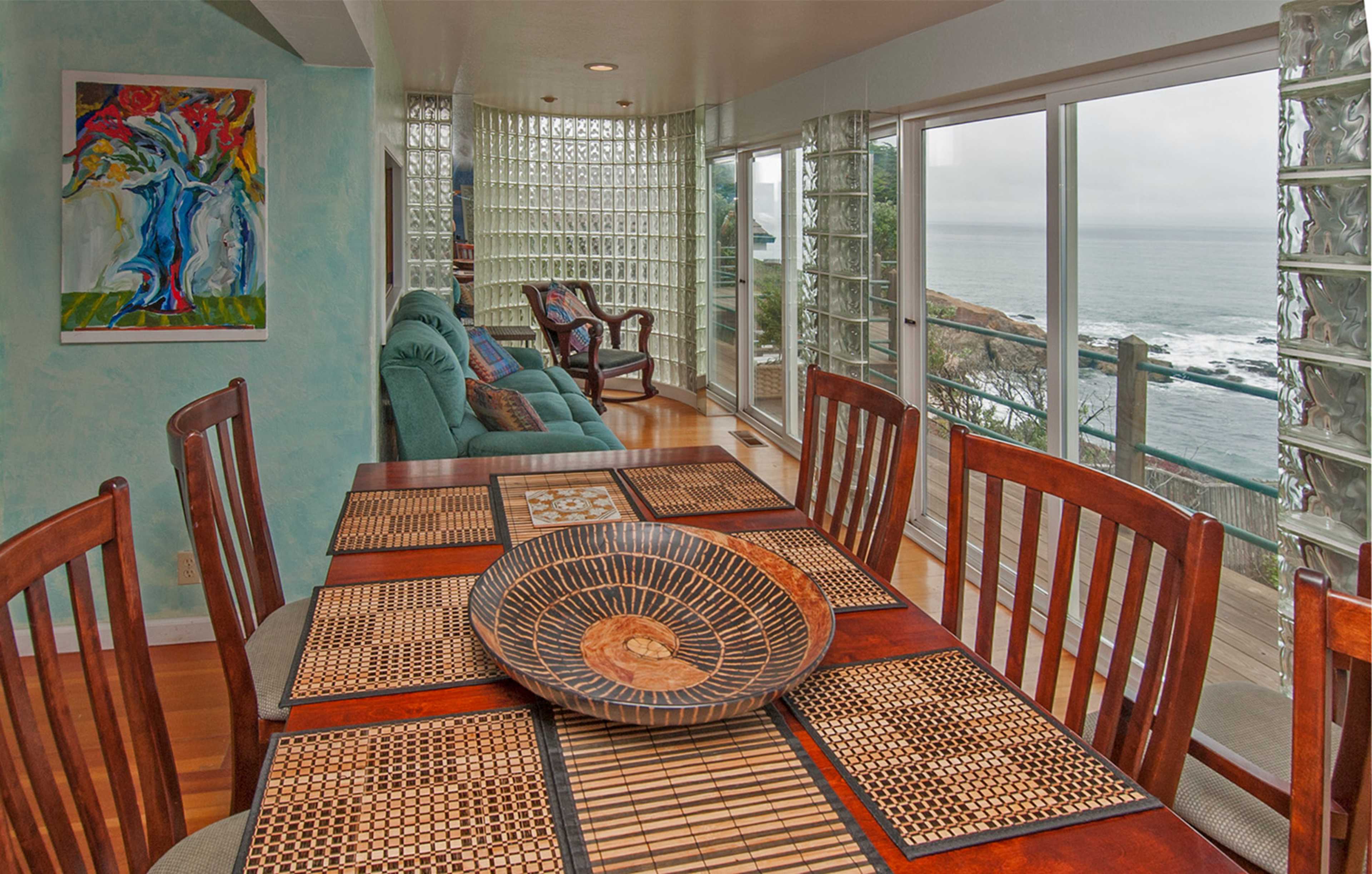 A dining area with a wooden table set on a woven placemat, overlooking a living space and large windows that provide a view of the ocean and nearby coastline.