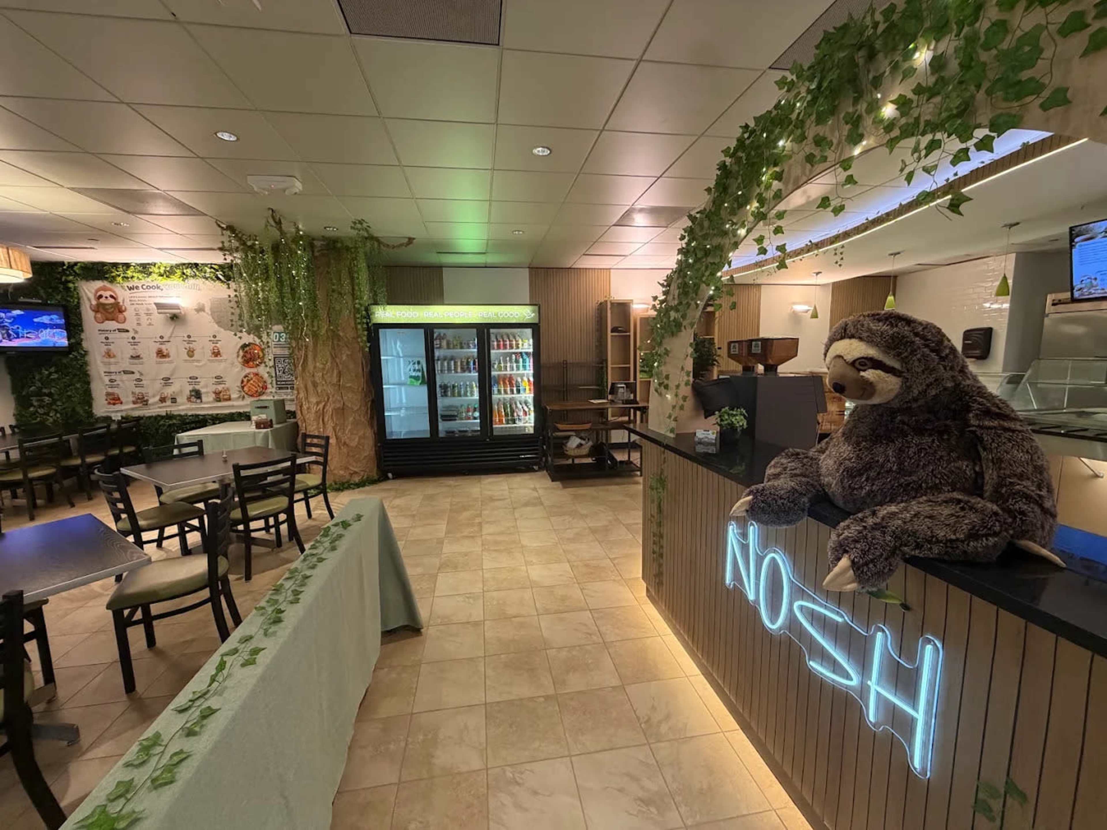 The image features a restaurant interior with a cozy ambiance, showcasing green plants along the walls, a display fridge, and a large plush sloth sitting on the counter.