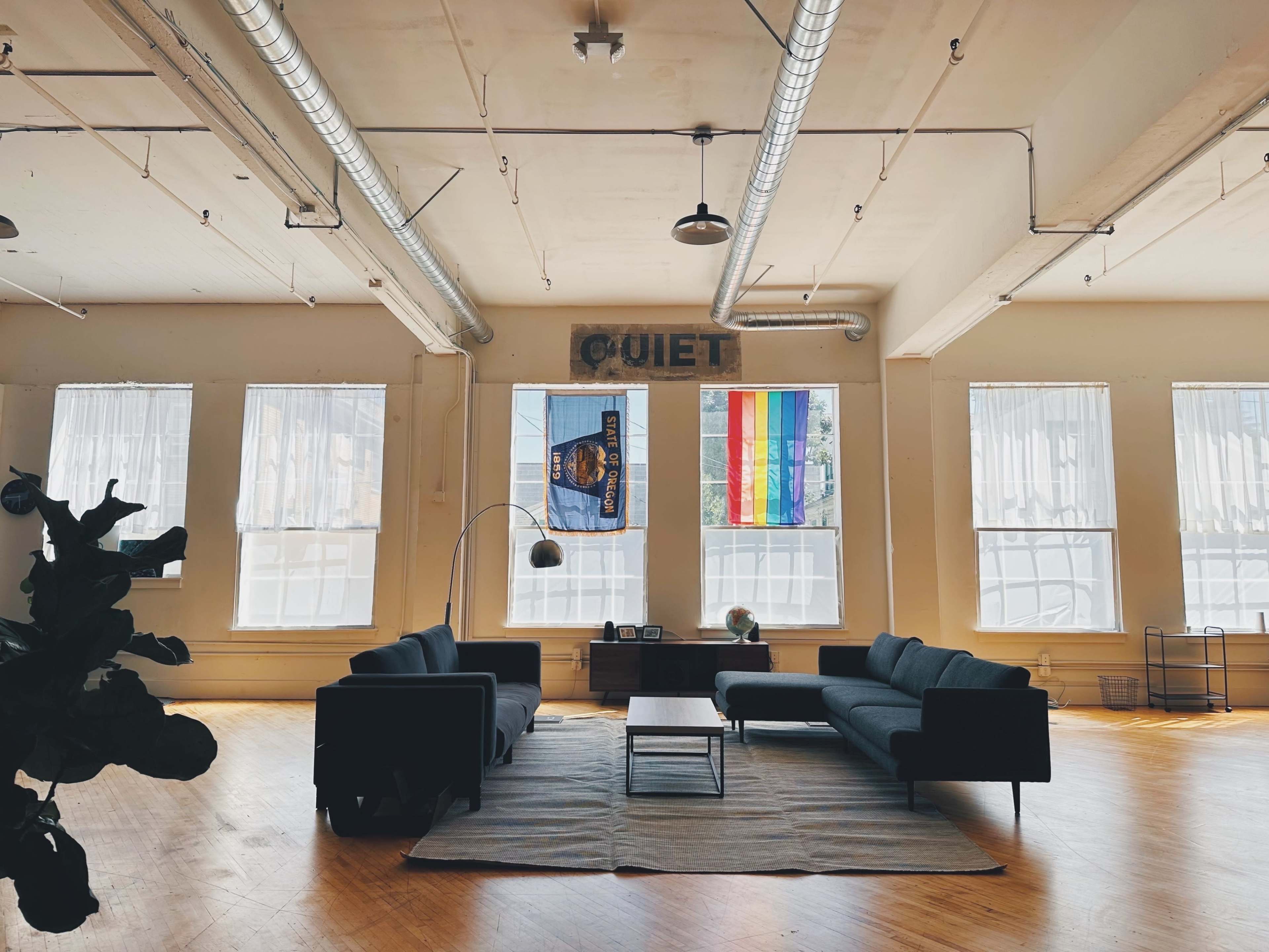 A spacious room with large windows, featuring a dark-colored sofa set, a rug on the wooden floor, and a prominent "QUIET" sign above the windows.