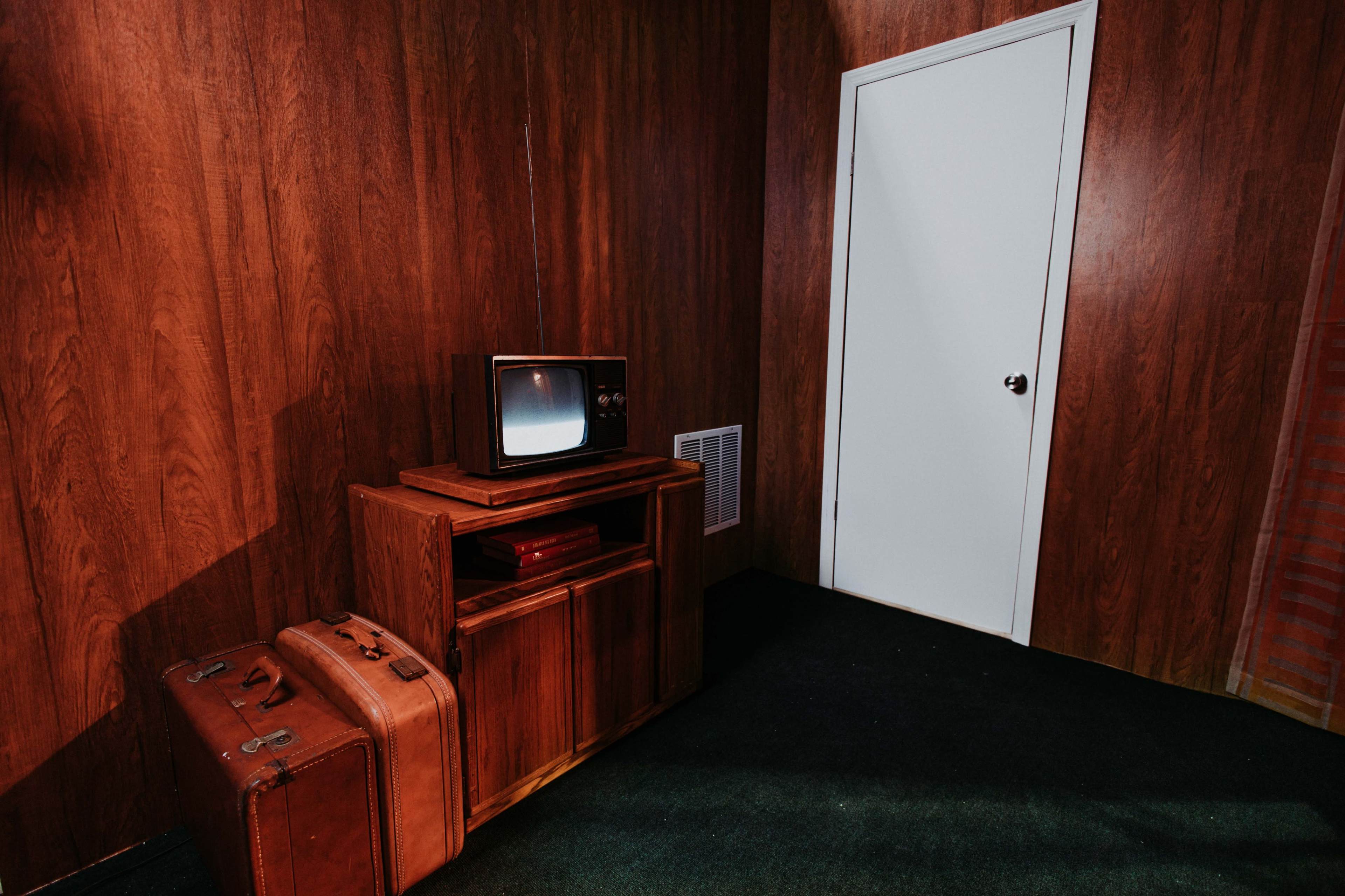 A small, wood-paneled room with a vintage television on a wooden stand next to two brown suitcases and a closed white door.
