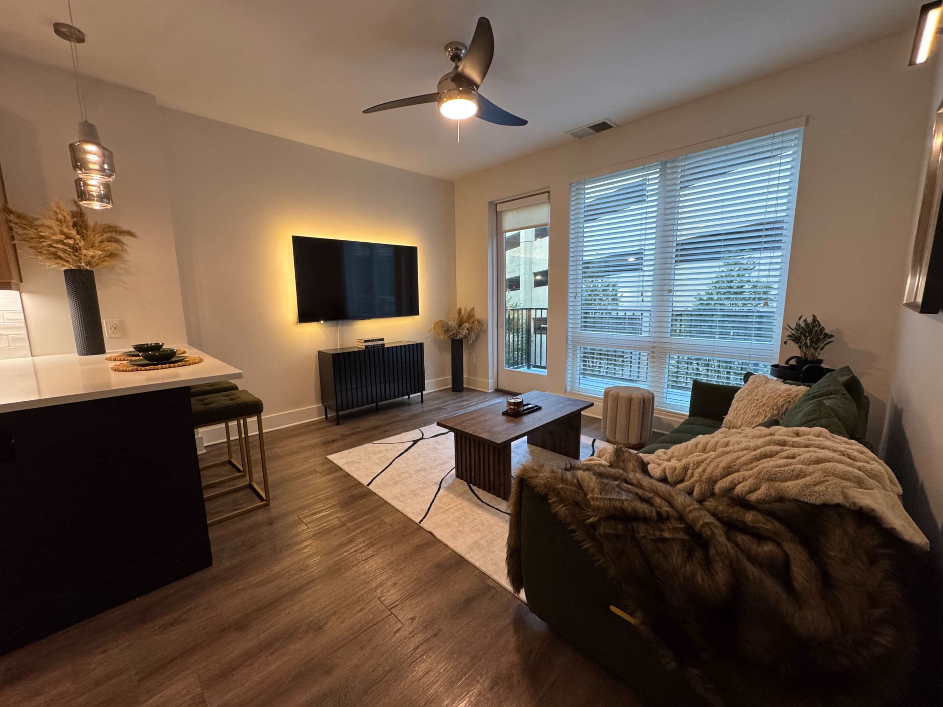 The room features a modern living area with a green sofa, a wooden coffee table, a television mounted on the wall, and large windows that allow natural light to enter.