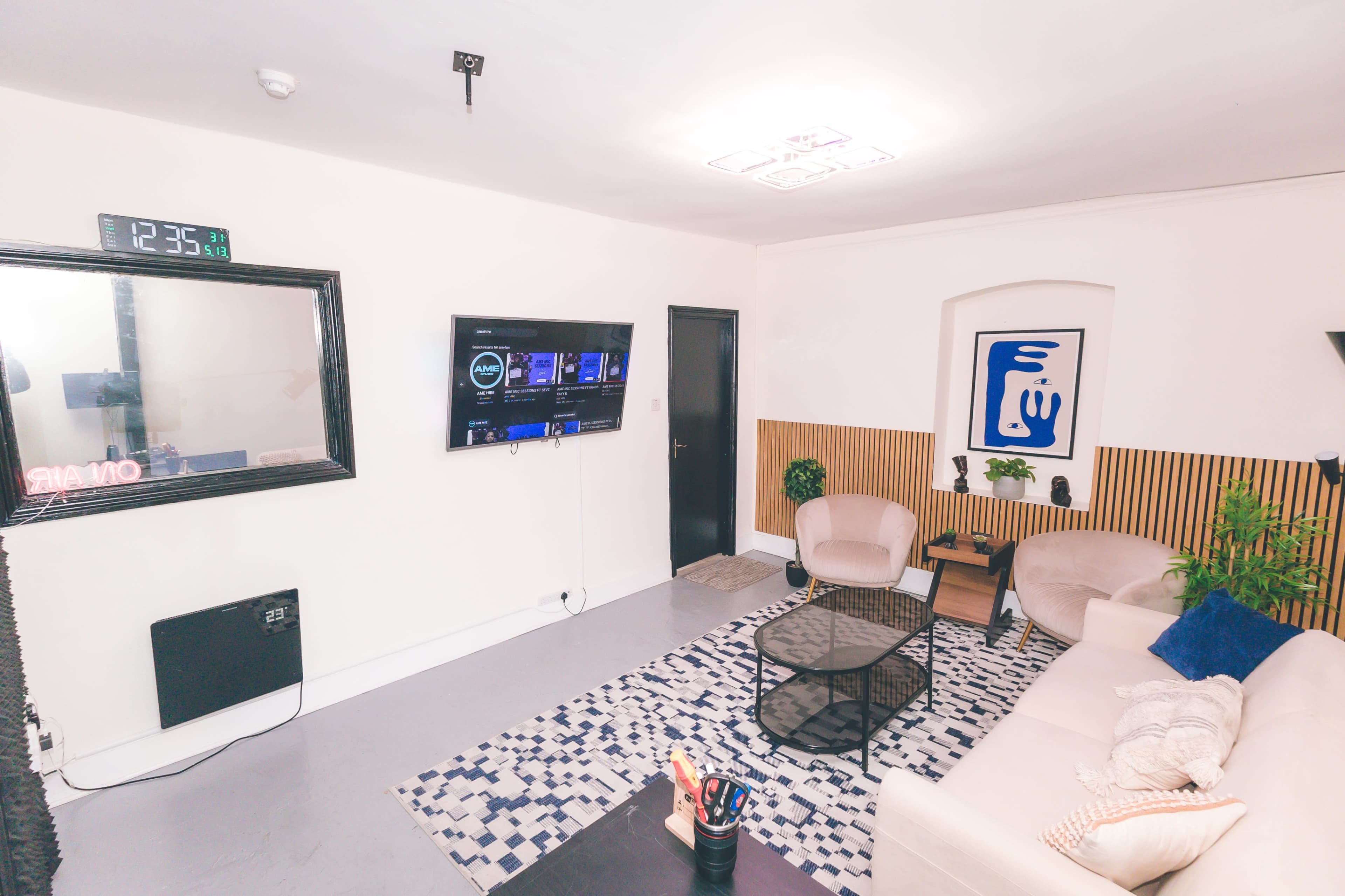 The image shows a modern lounge area with two armchairs, a sofa, a coffee table, a television mounted on the wall, and decorative plants.
