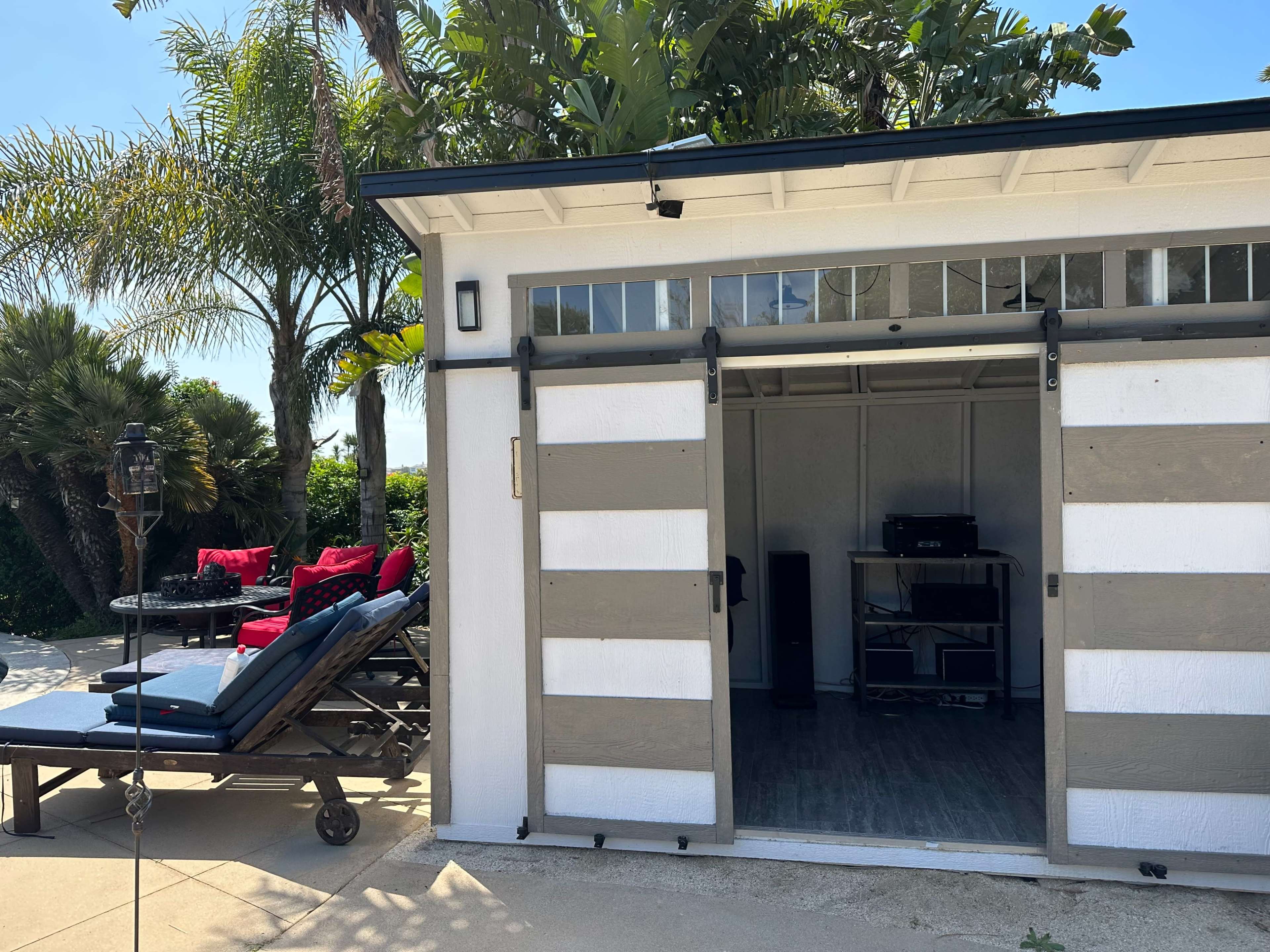 A shed with sliding doors opens to reveal a small audio setup, surrounded by a patio area featuring lounge chairs and a table with red cushions.